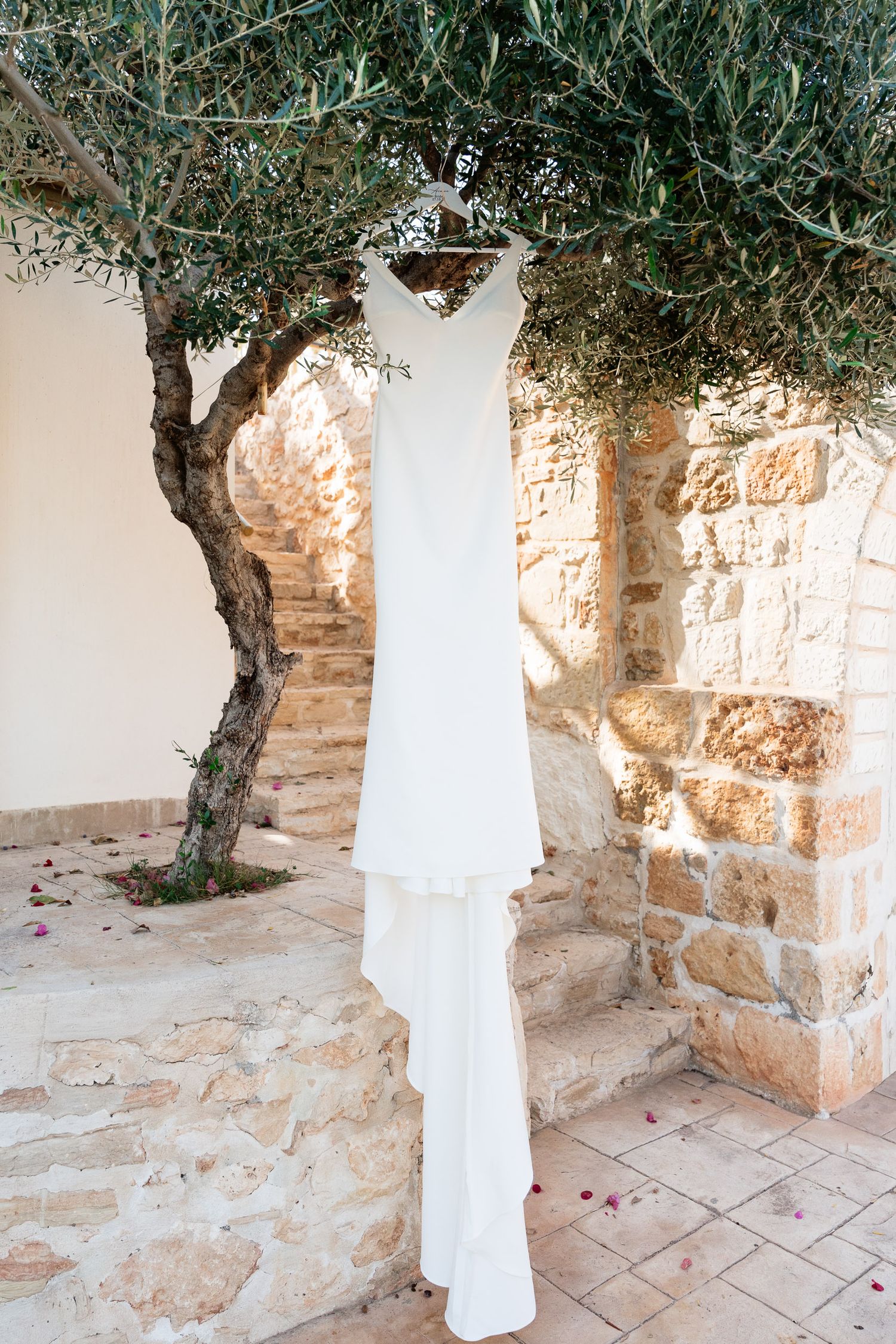 A white wedding dress hangs elegantly against a rustic stone wall backdrop with an olive tree and scattered rose petals.