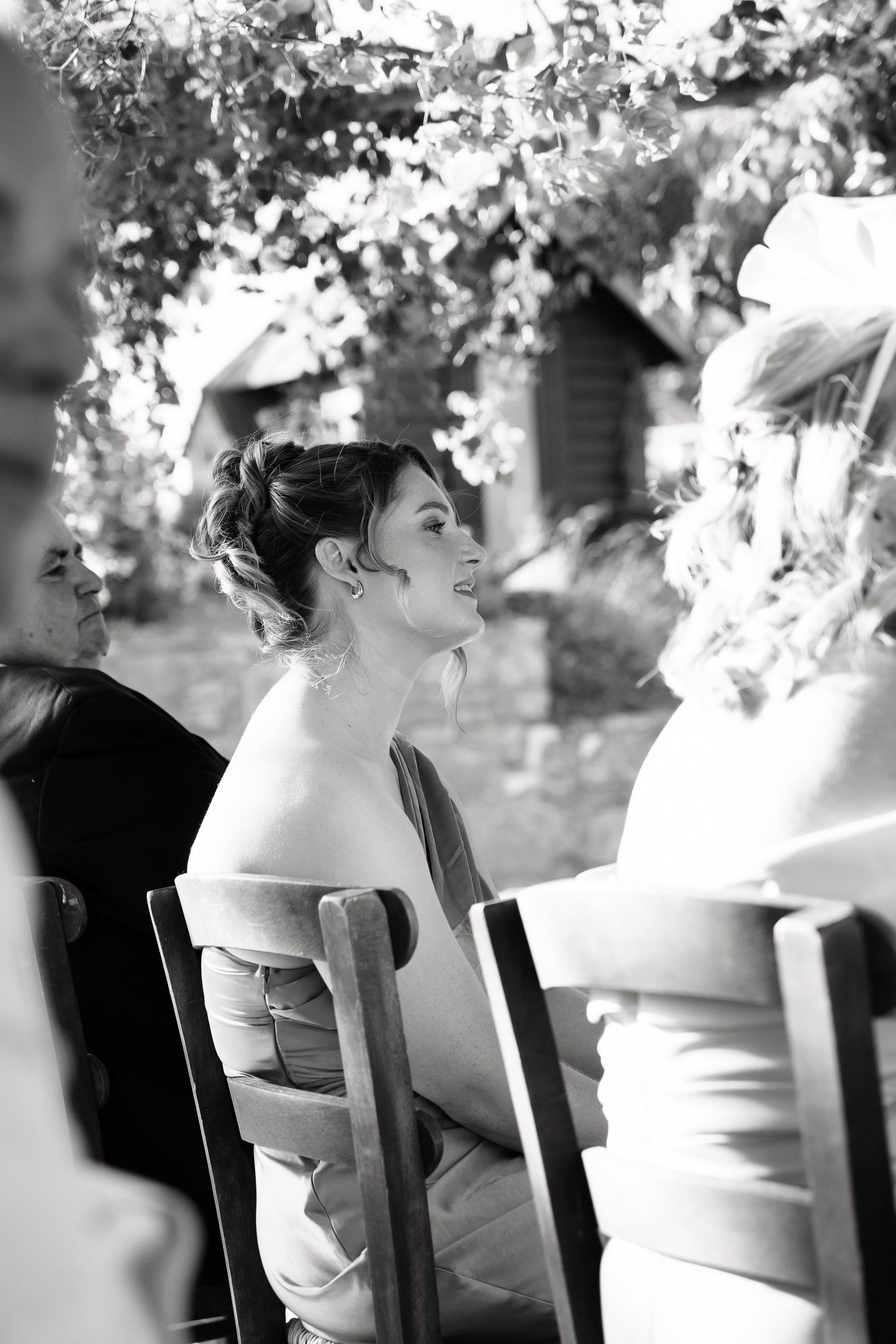Black and white candid photograph of wedding guests seated in wooden chairs during an outdoor garden reception.