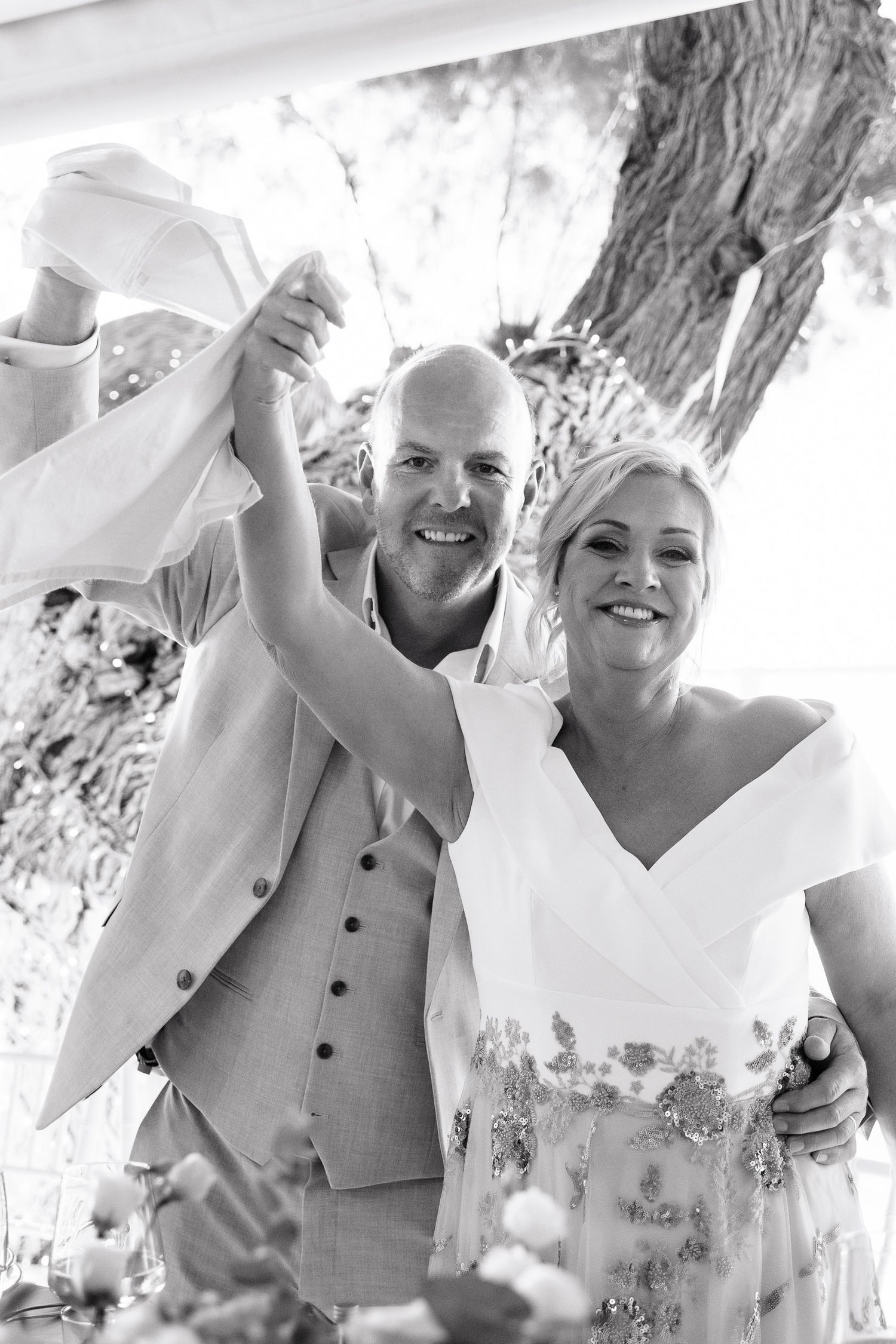 A joyful wedding party in light colored attire laughs together in an outdoor setting.