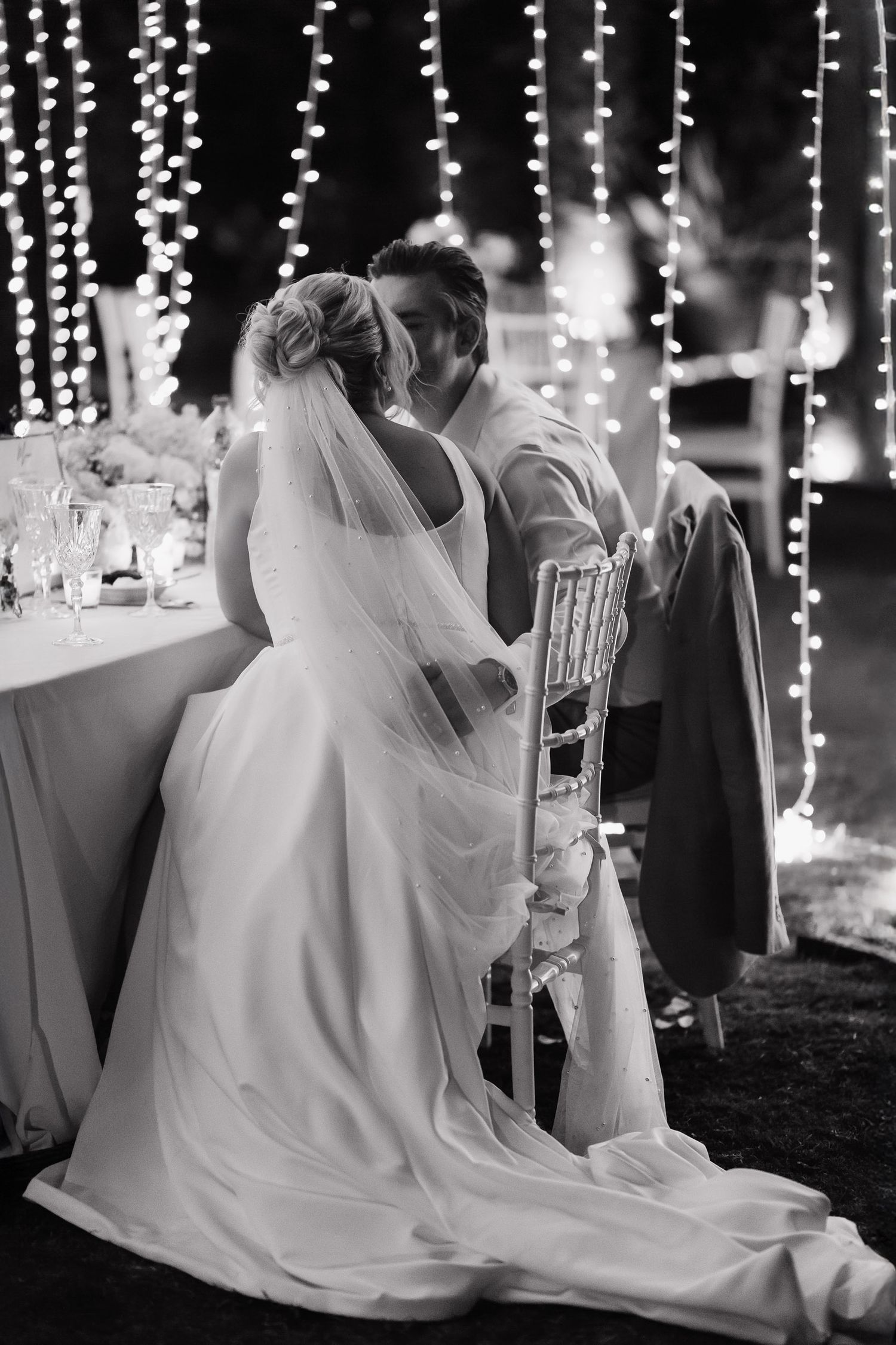 A romantic black and white wedding moment captured under twinkling string lights with a flowing white dress and veil.