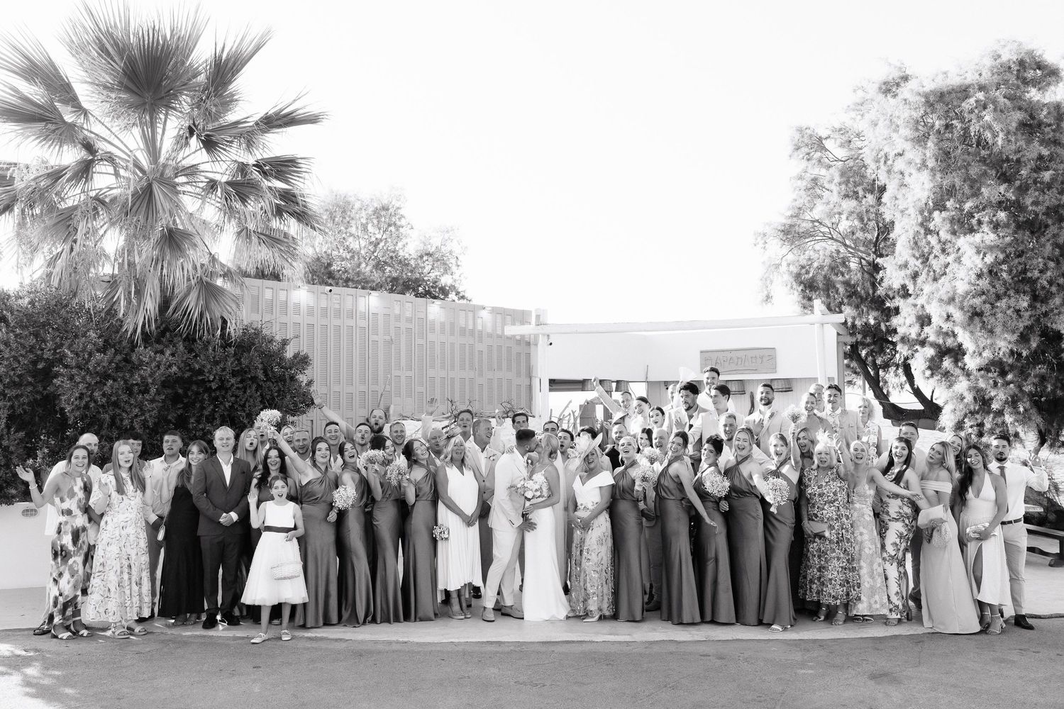 A black and white panoramic photo shows a large wedding party gathered outdoors with palm trees in the background.