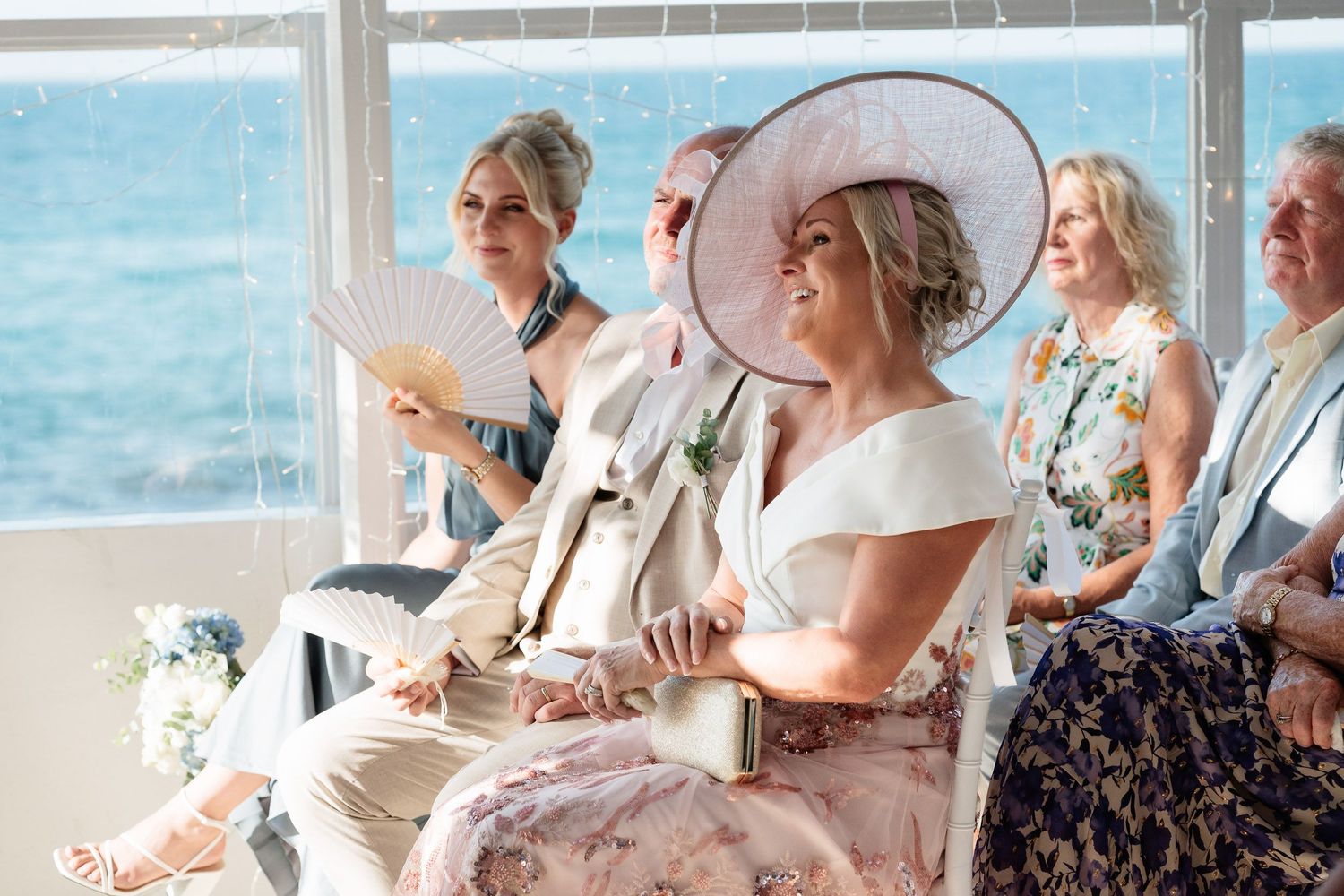 Wedding guests enjoying a beachfront celebration with white decor and floral arrangements by the ocean.