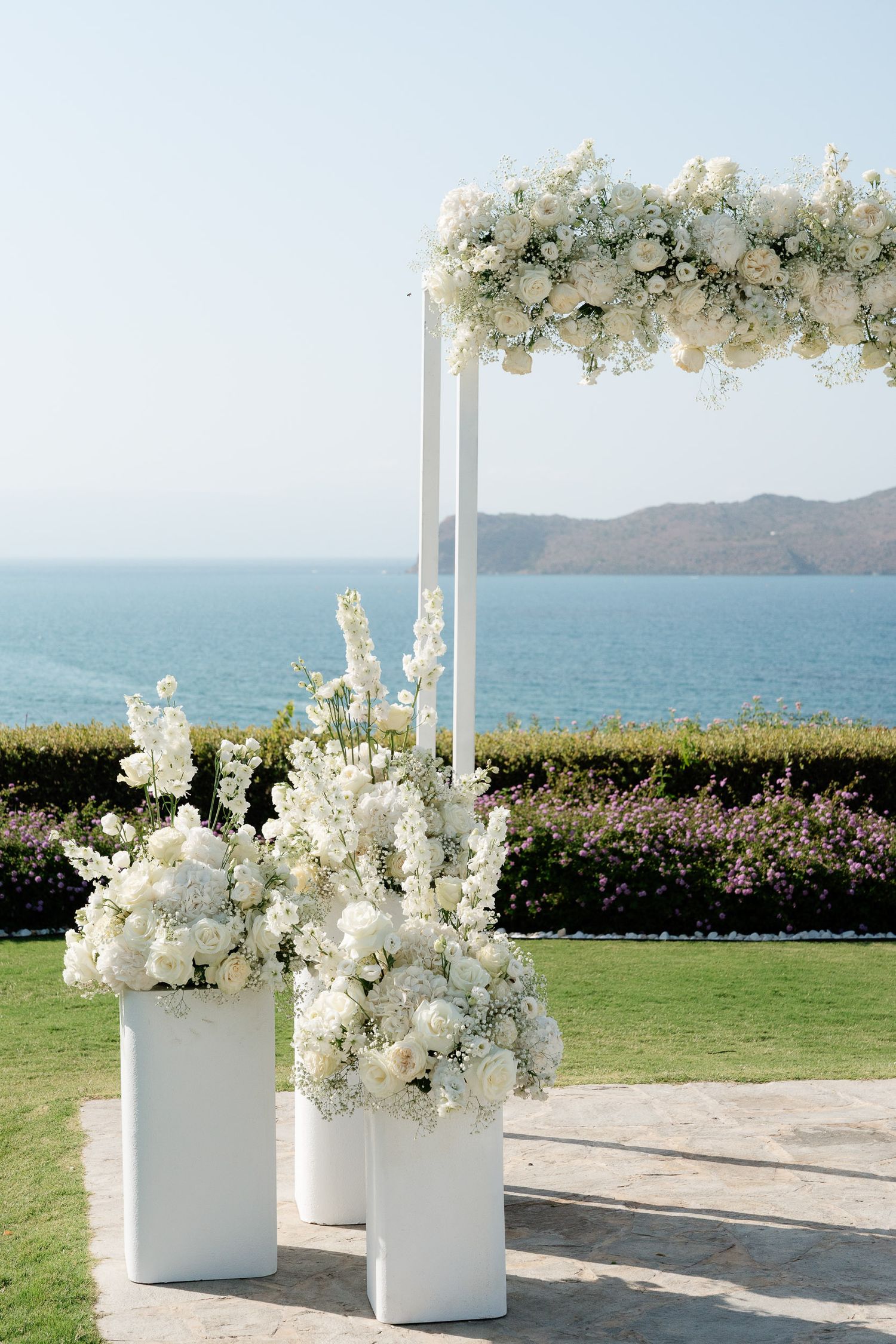 White floral arrangements in elegant columns create a romantic seaside wedding ceremony setup overlooking the ocean.