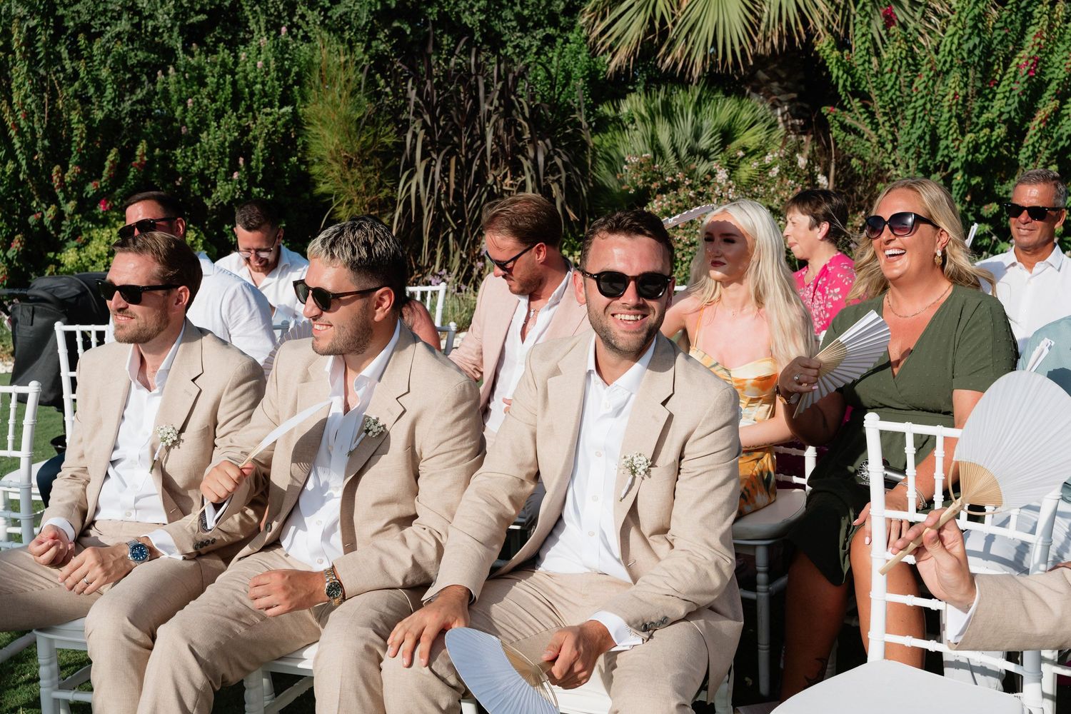 Groomsmen in tan suits share laughter and conversation while sitting in white chairs at an outdoor wedding venue.