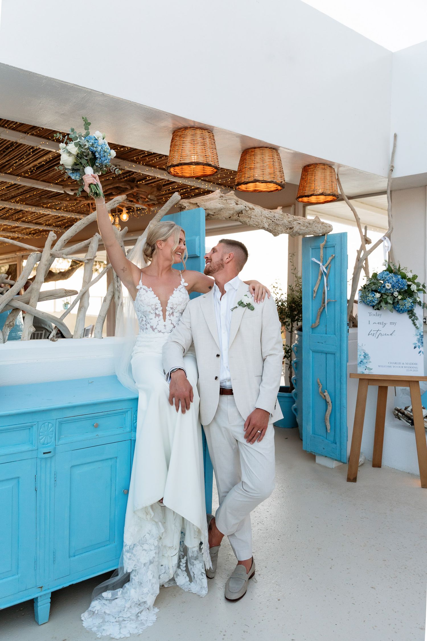 Wedding couple poses against bright turquoise doors and white walls in a rustic indoor setting with woven lamp shades.