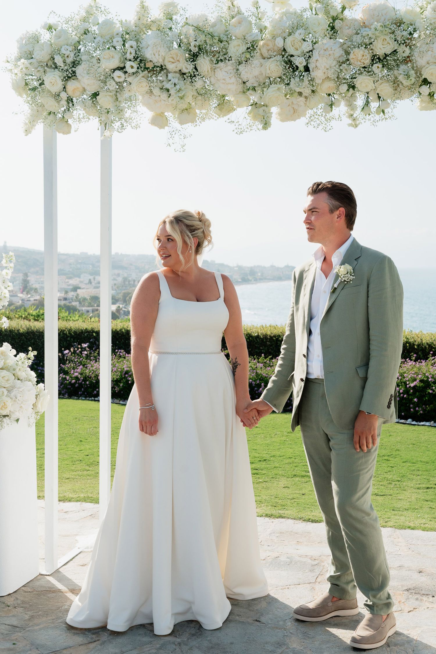 A couple in wedding attire shares tender moments under a white floral arch at an outdoor venue with scenic views.