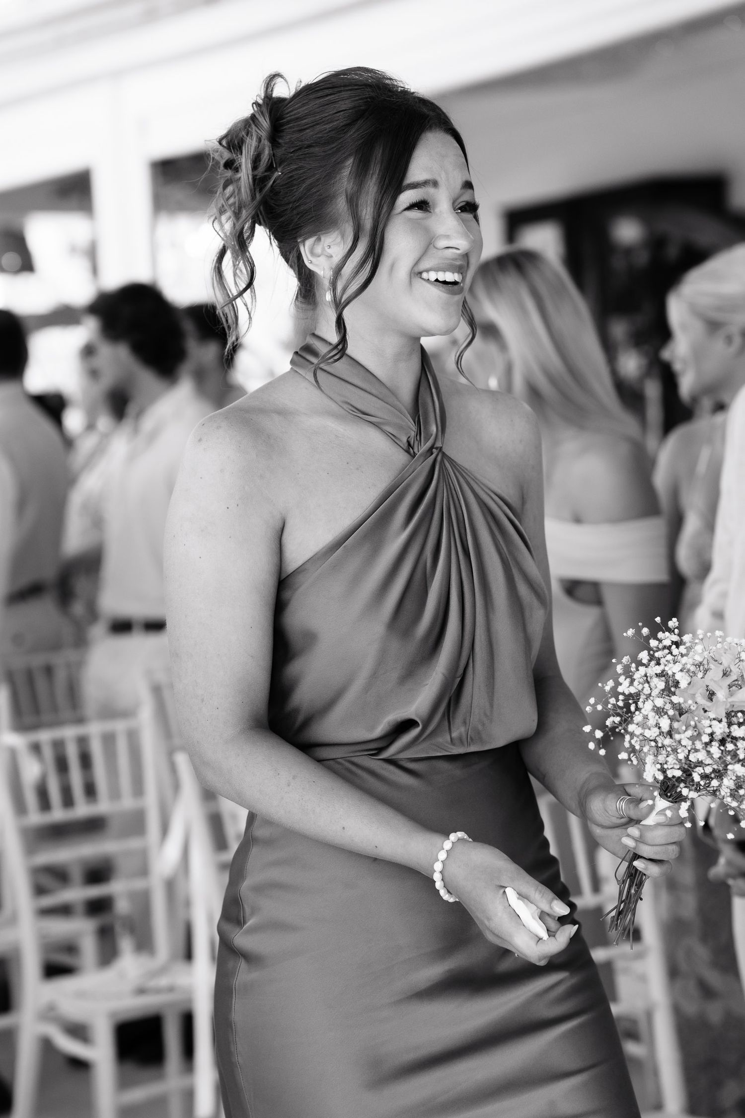 A bridesmaid in an elegant halter dress smiles joyfully while holding a small bouquet at a wedding celebration.