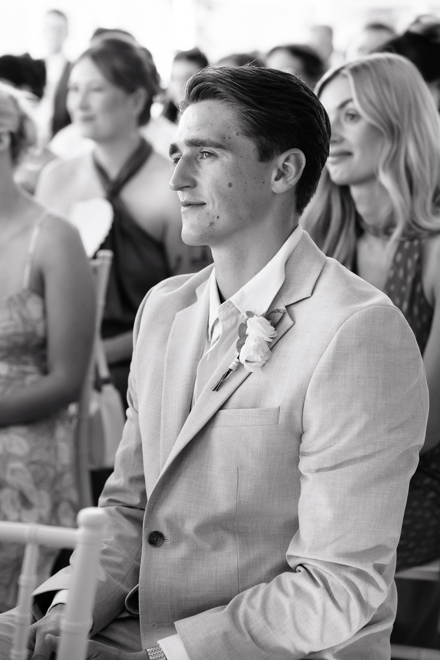 A black and white photo shows groomsmen in light colored suits watching attentively during a wedding ceremony.
