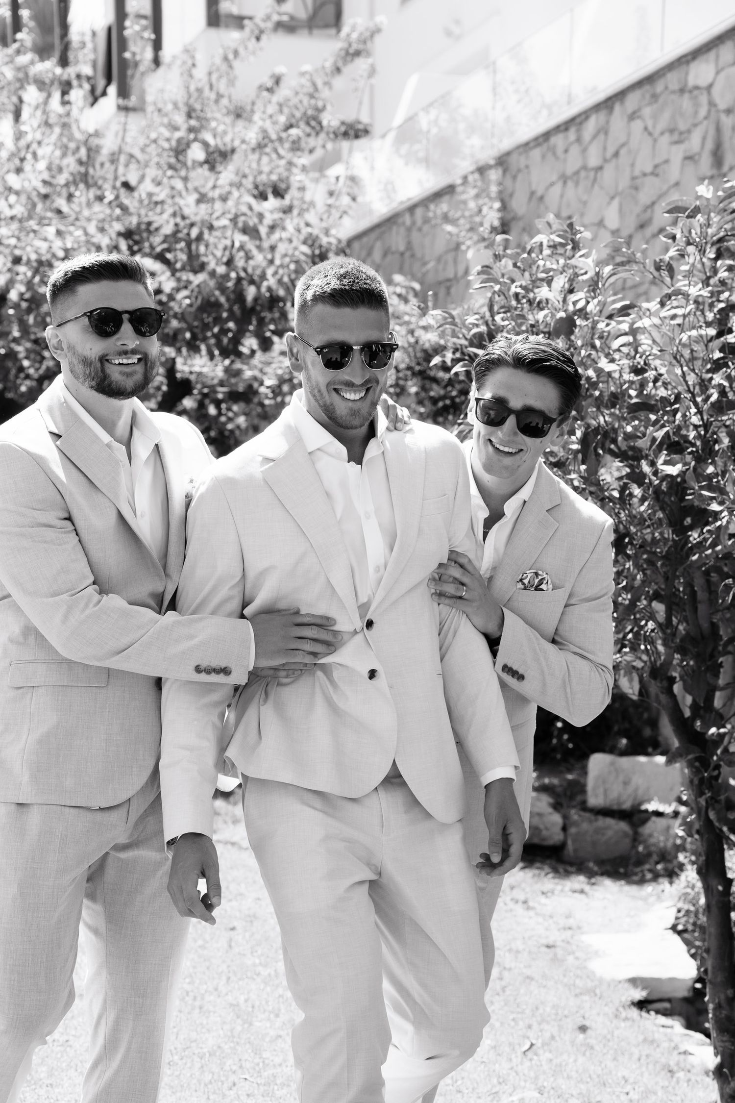Three people wearing white suits and sunglasses pose playfully together outdoors on a sunny day.