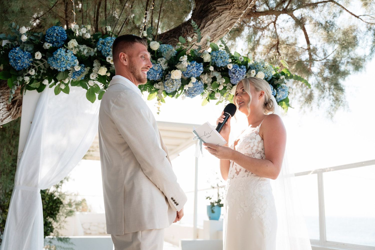 A couple exchanges vows beneath a floral arch decorated with blue flowers during an outdoor wedding ceremony.