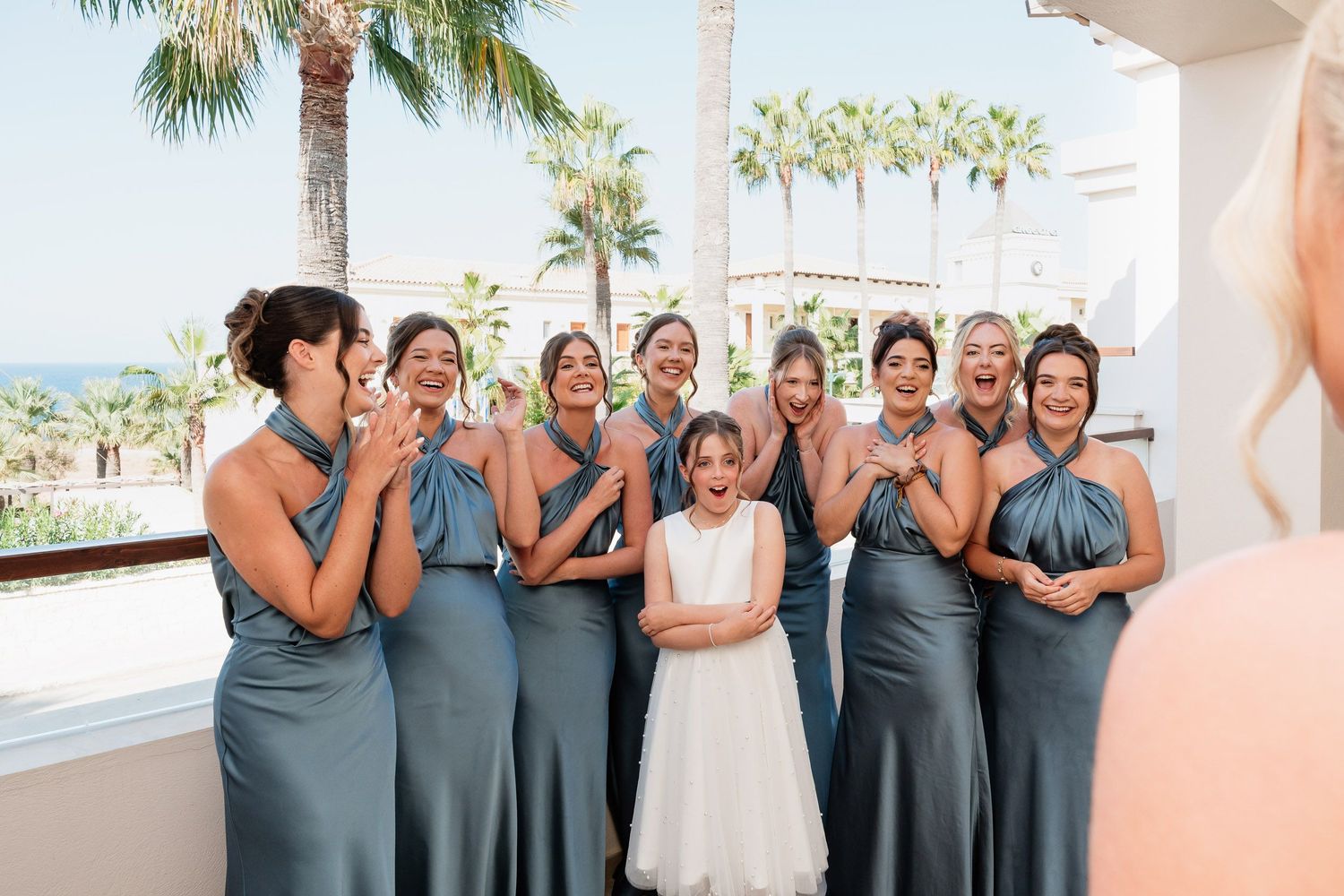 Wedding party in coordinated blue dresses poses with the bride in white against a tropical palm tree backdrop.