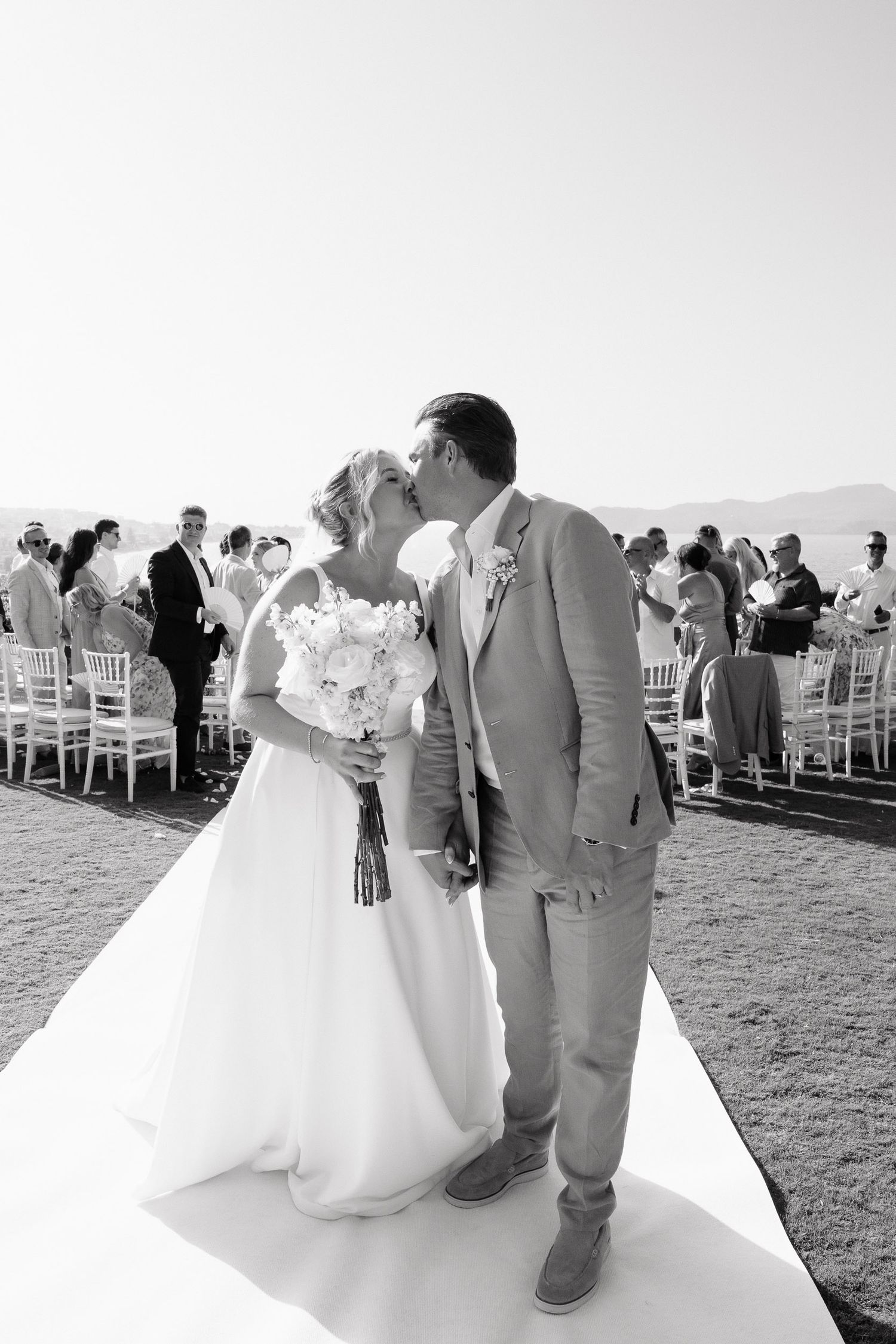 A black and white panoramic sequence of a wedding ceremony on a grassy hilltop with guests seated in white chairs.