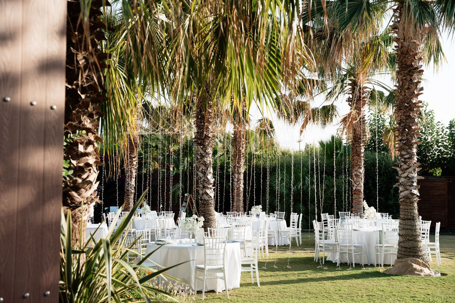 Outdoor wedding venue with white chairs arranged in rows under palm trees and string lights at dusk.