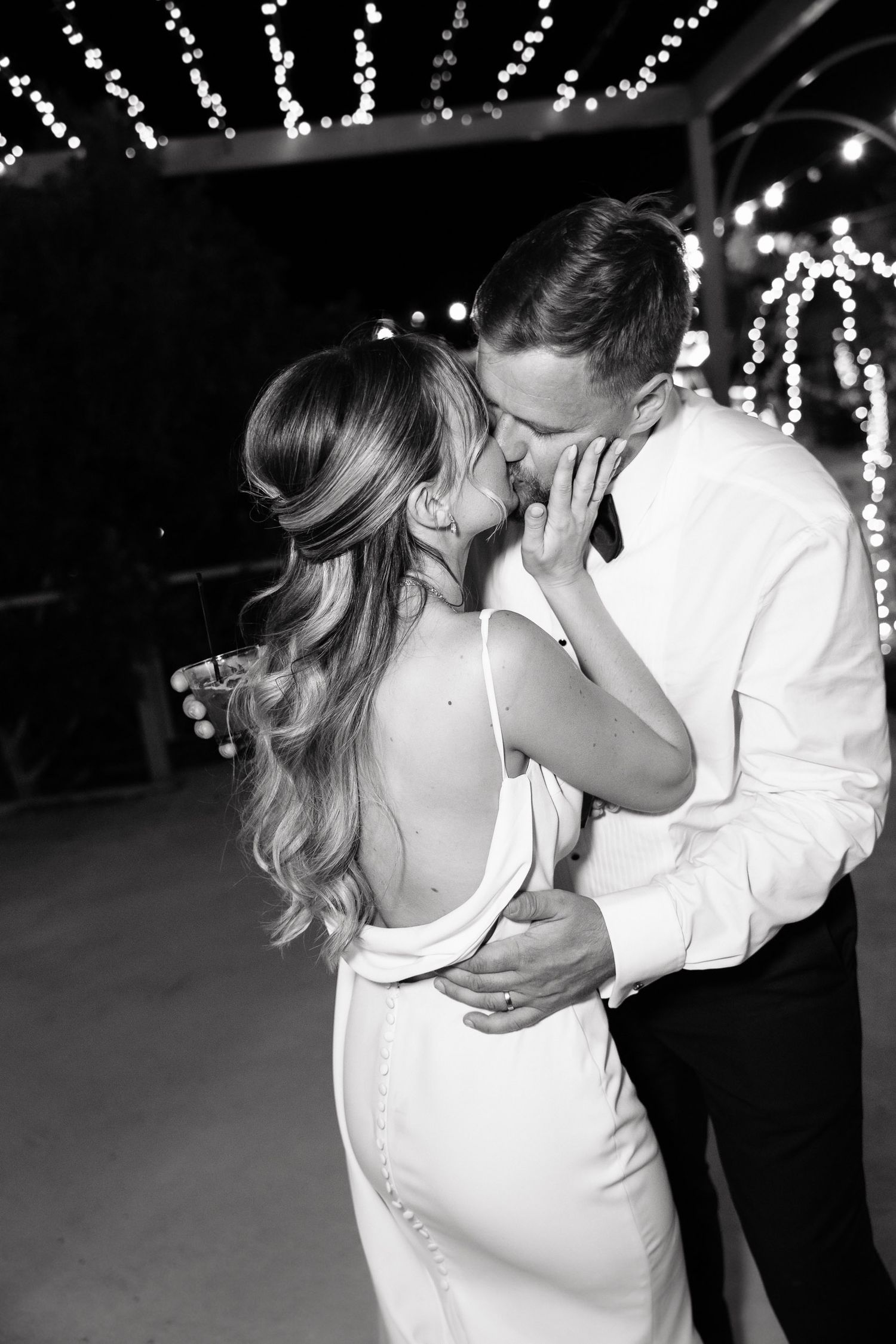 A couple in white attire dancing together at a nighttime wedding reception with twinkling lights in the background.