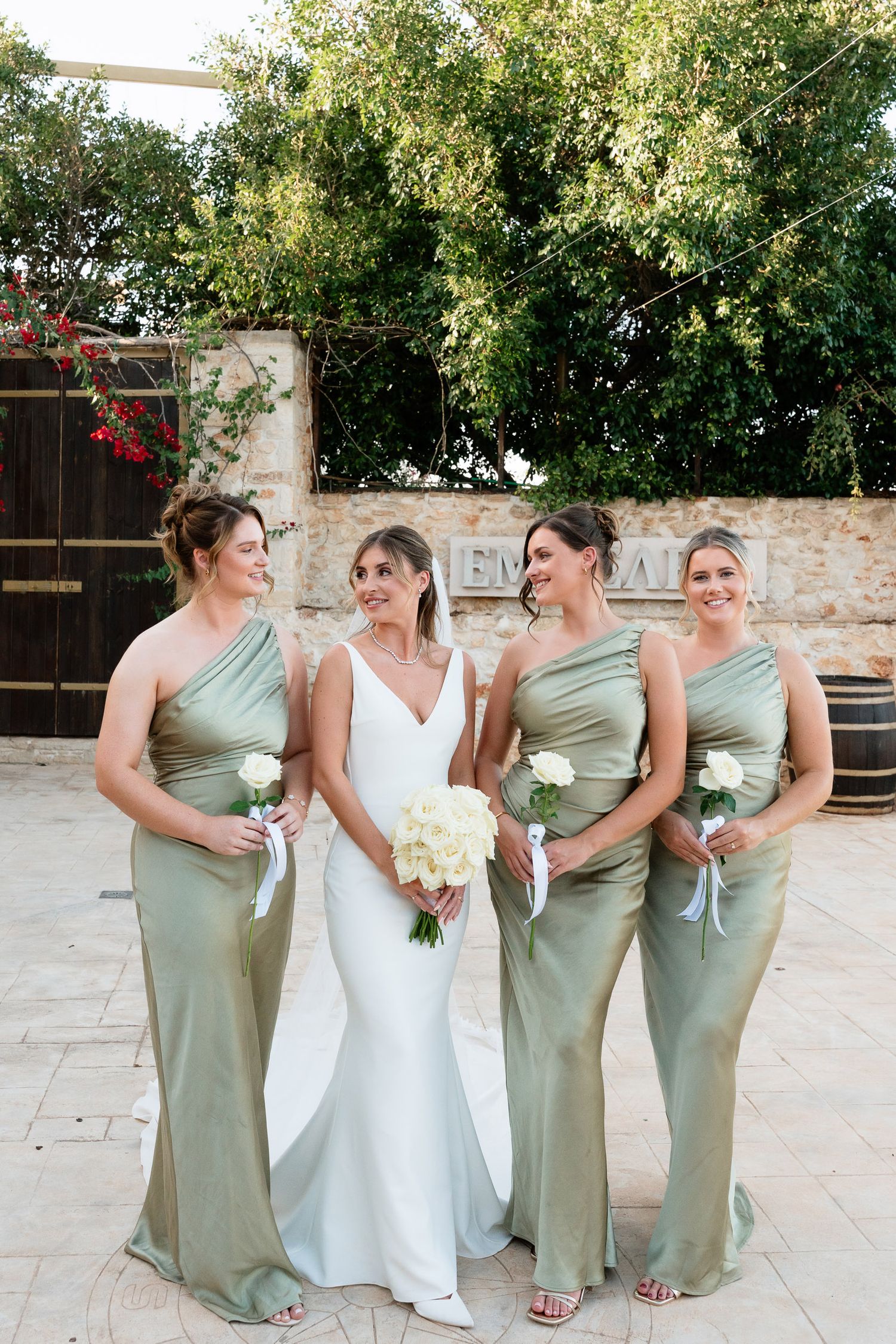 Wedding party in sage green dresses and white gown pose together on stone patio during outdoor celebration.