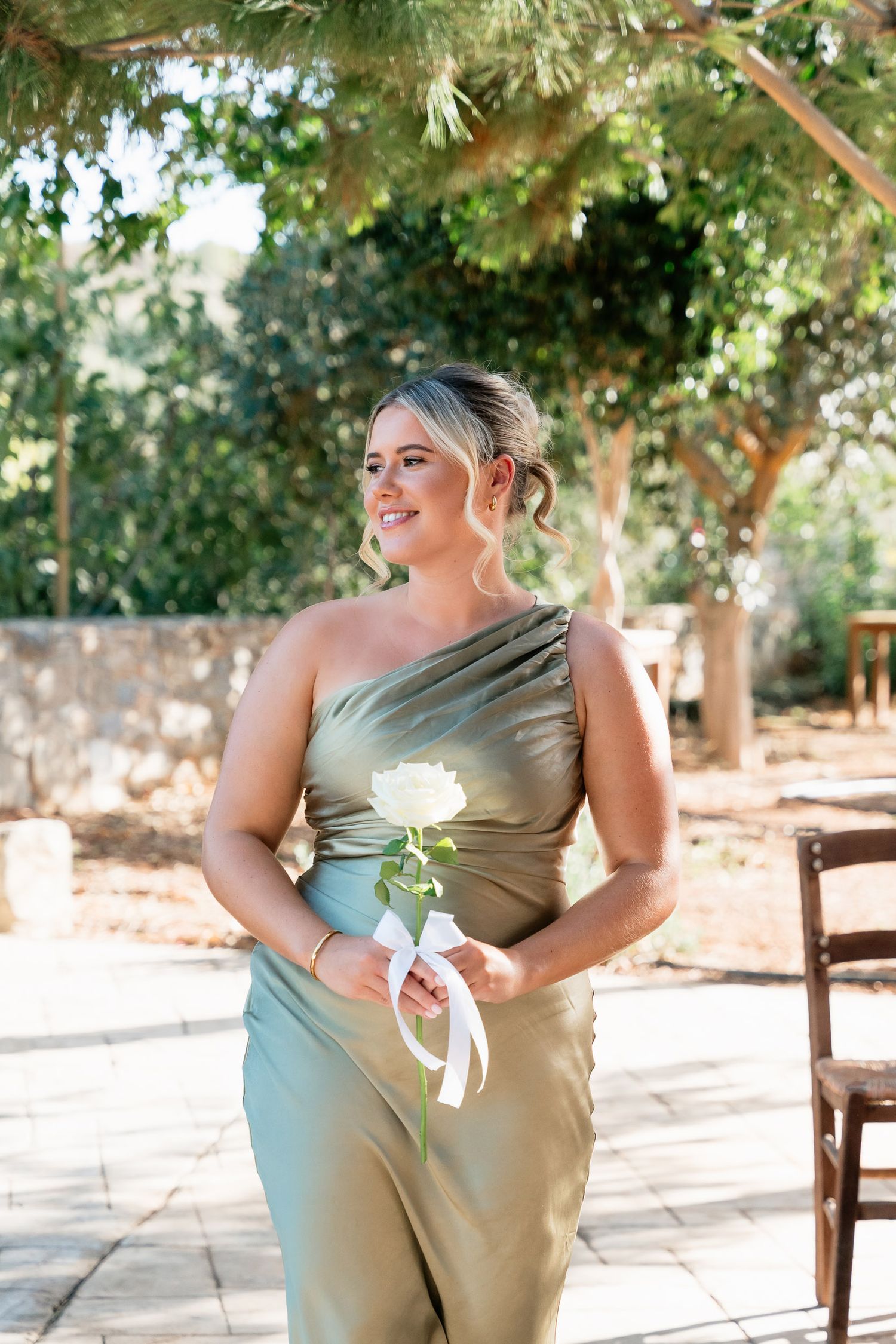 A bridesmaid in a sage green one-shoulder dress holds a white rose while standing outdoors in natural lighting.