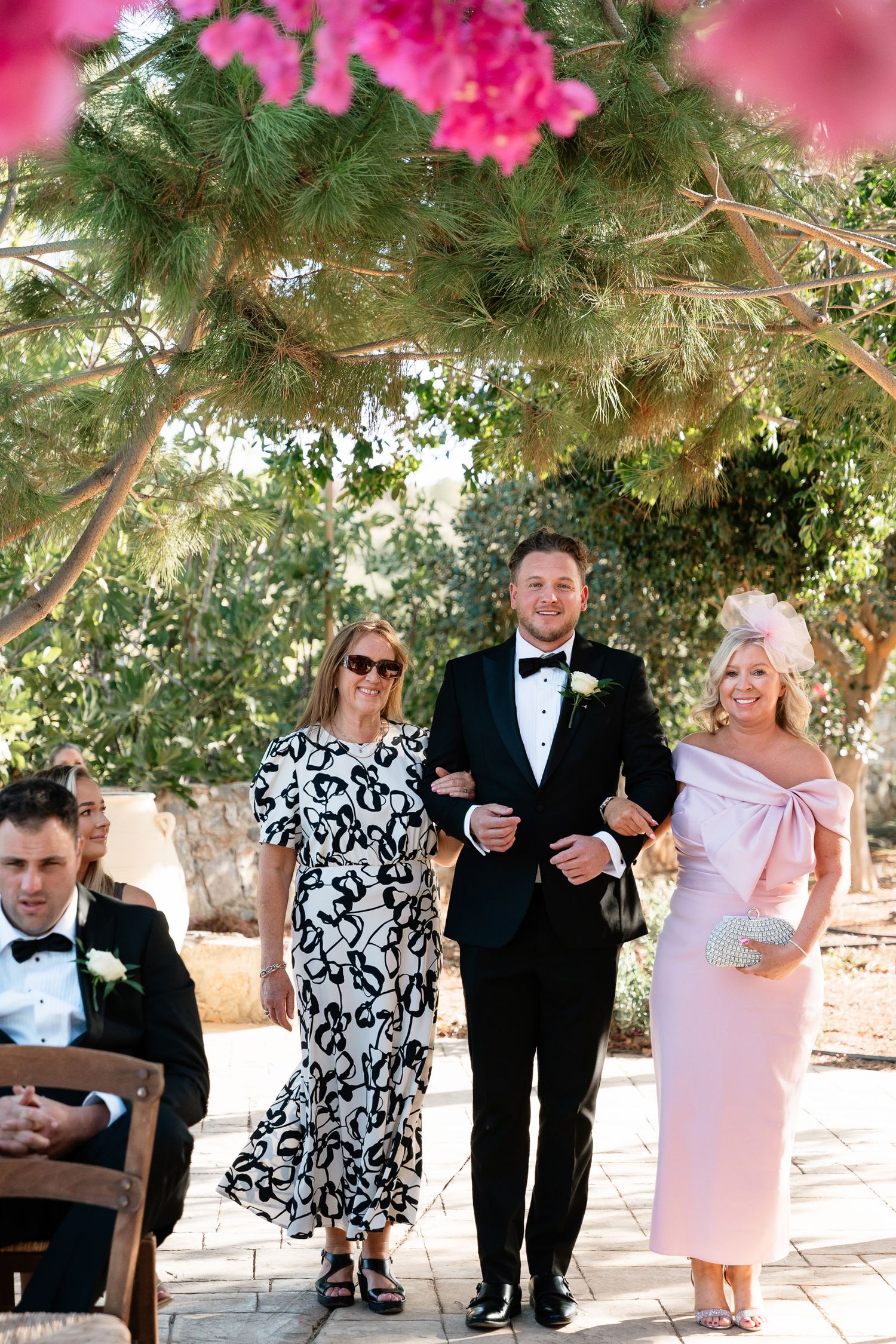 A wedding party walks along a garden path under trees with vibrant pink flowers blooming overhead.