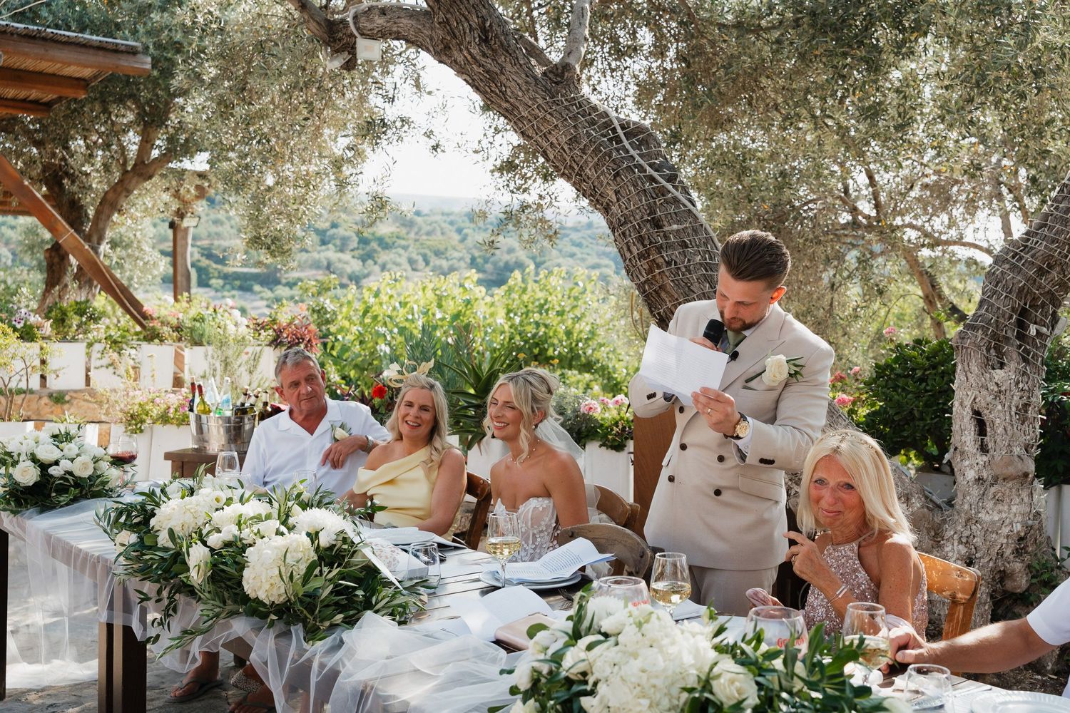 Wedding guests enjoying an outdoor reception dinner under olive trees in a Mediterranean setting.