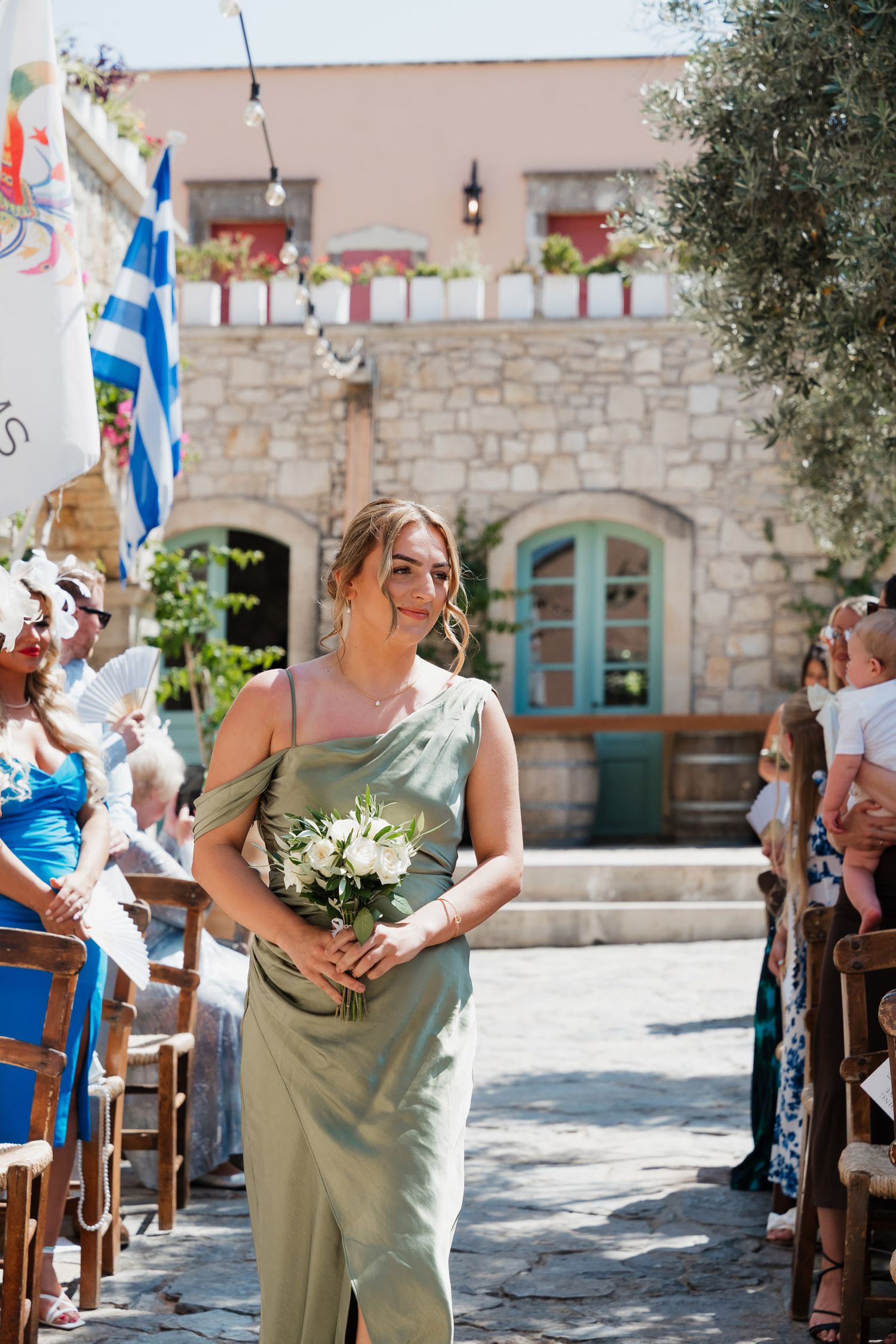 Wedding party members in sage green dresses celebrate outside a Mediterranean stone building with flags and decorations.