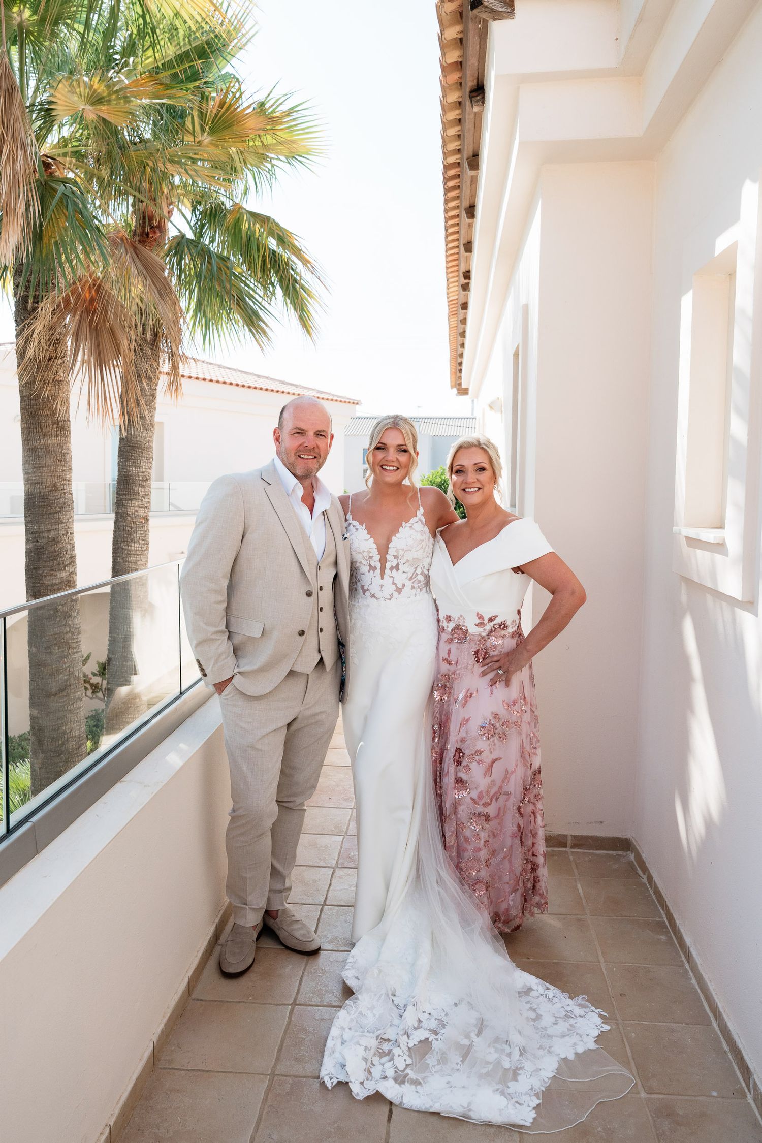 Three people pose together on a Mediterranean-style balcony with palm trees and white stucco walls on a sunny day.