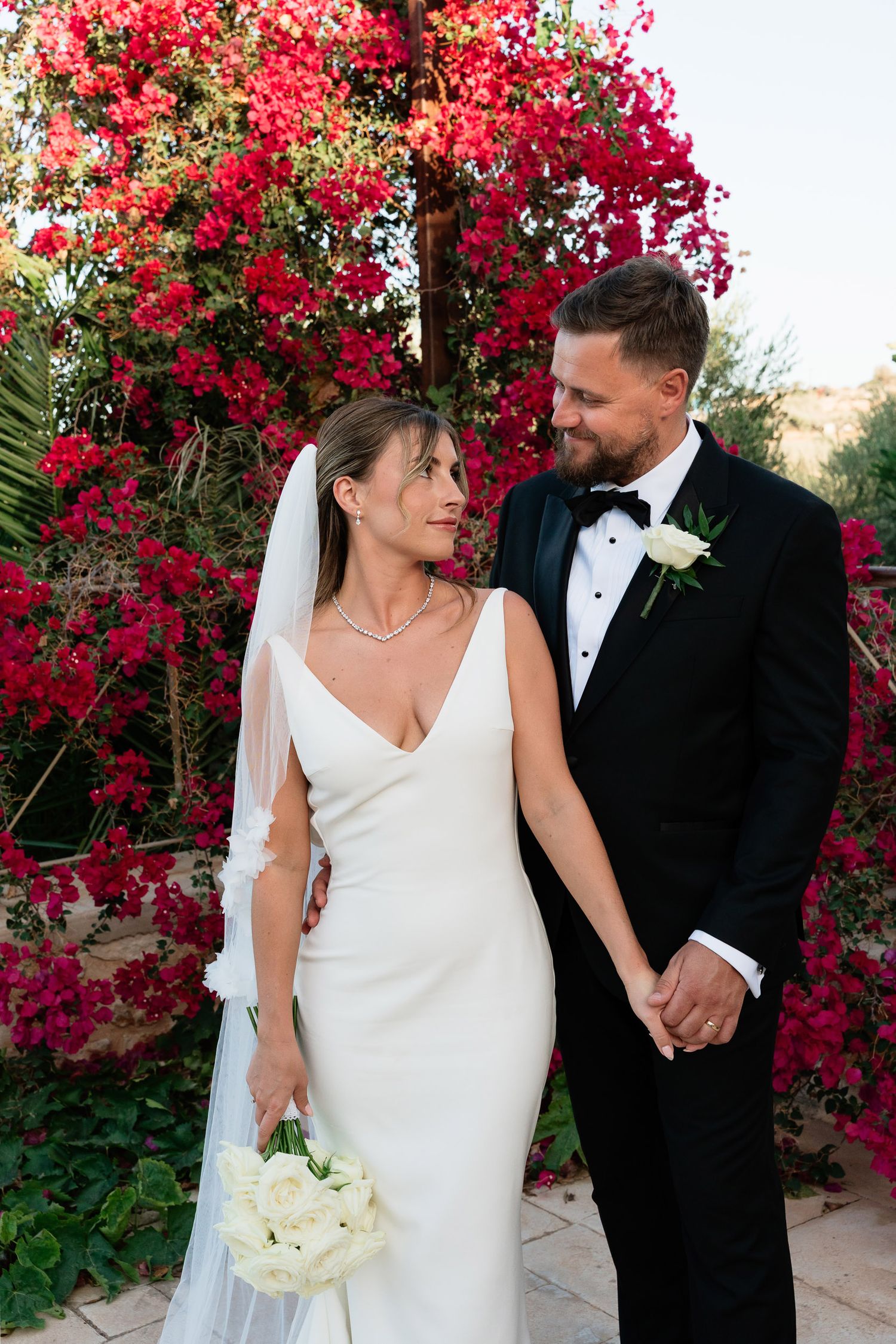 A romantic wedding couple poses against vibrant red bougainvillea flowers in an outdoor garden setting.
