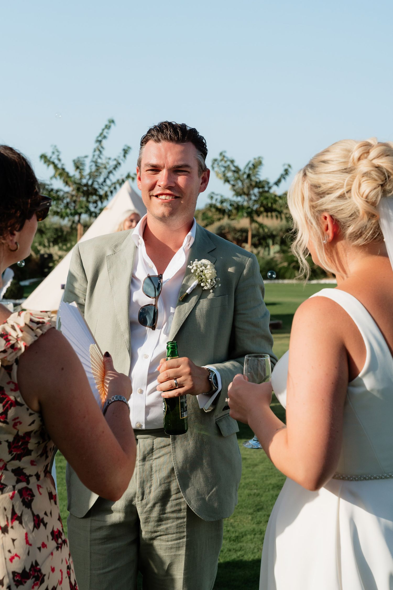 Wedding guests enjoying drinks and conversation on a sunny day at an outdoor reception on a grassy lawn.
