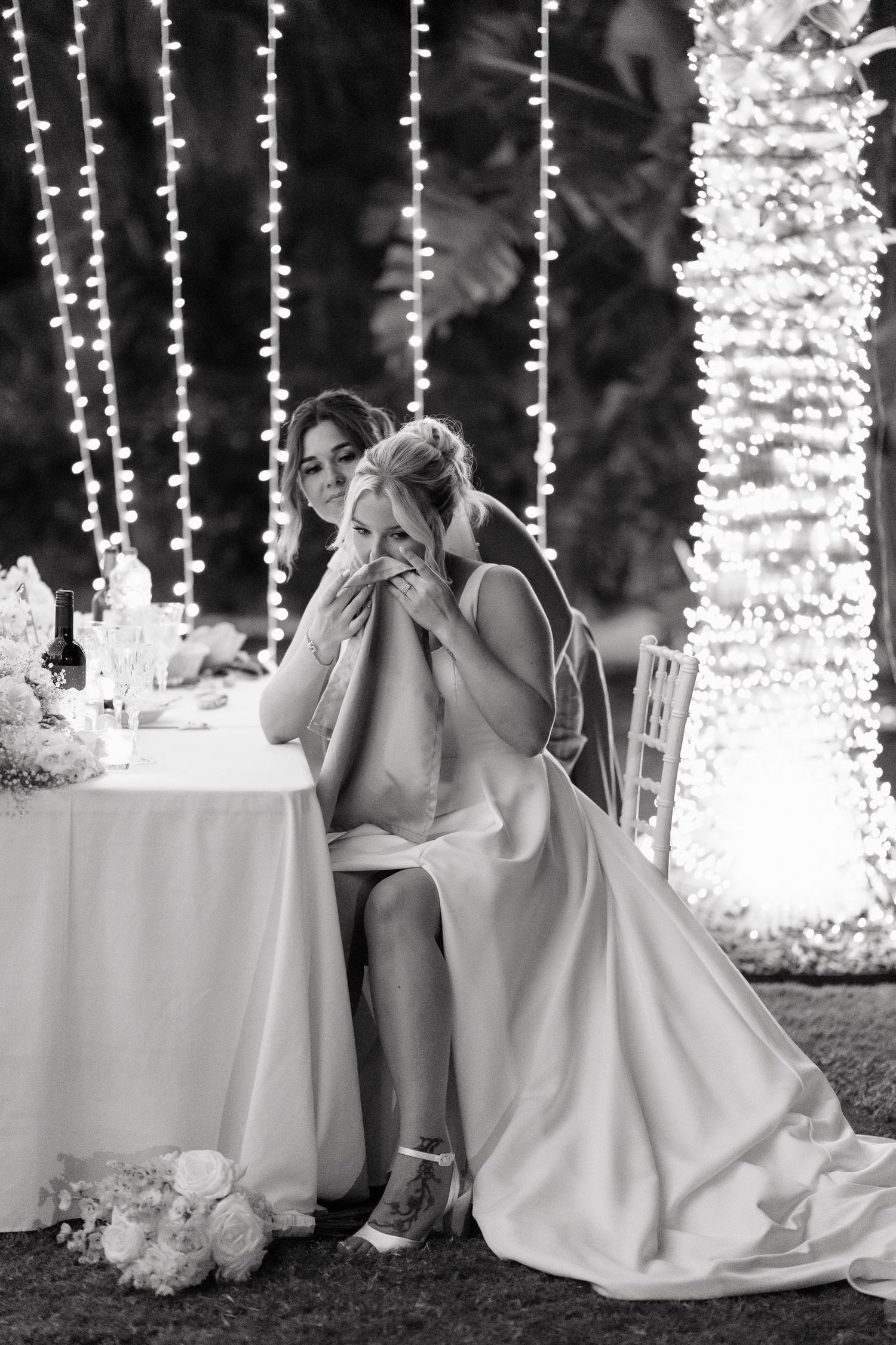 Black and white wedding photo showing an emotional moment at a reception with twinkle lights and floral decorations.