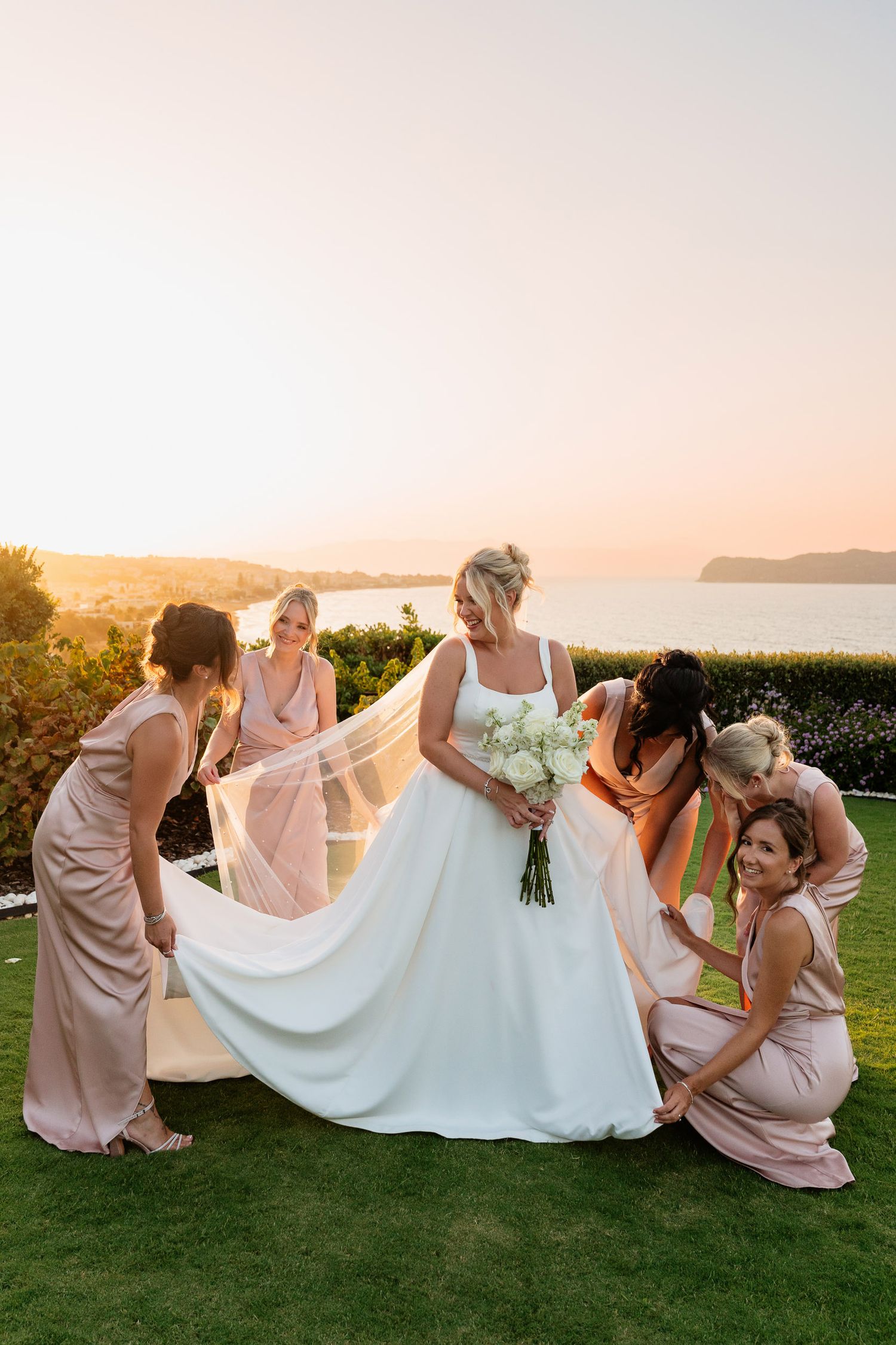 A wedding party in blush pink dresses poses playfully with the bride's white gown on a grassy lawn at sunset.
