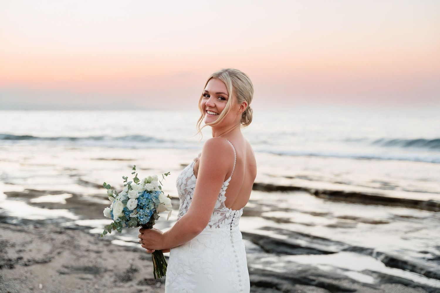 A beaming bride holds a blue and white bouquet while standing on a beach at sunset with ocean waves in the background.