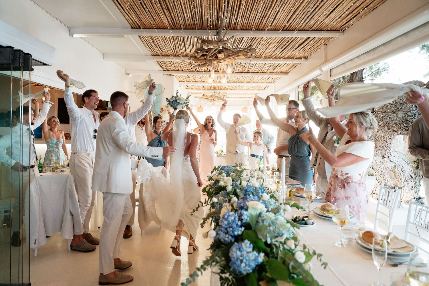 Wedding reception guests in white attire celebrate with raised glasses at a bright coastal venue decorated with blue hydrangeas.