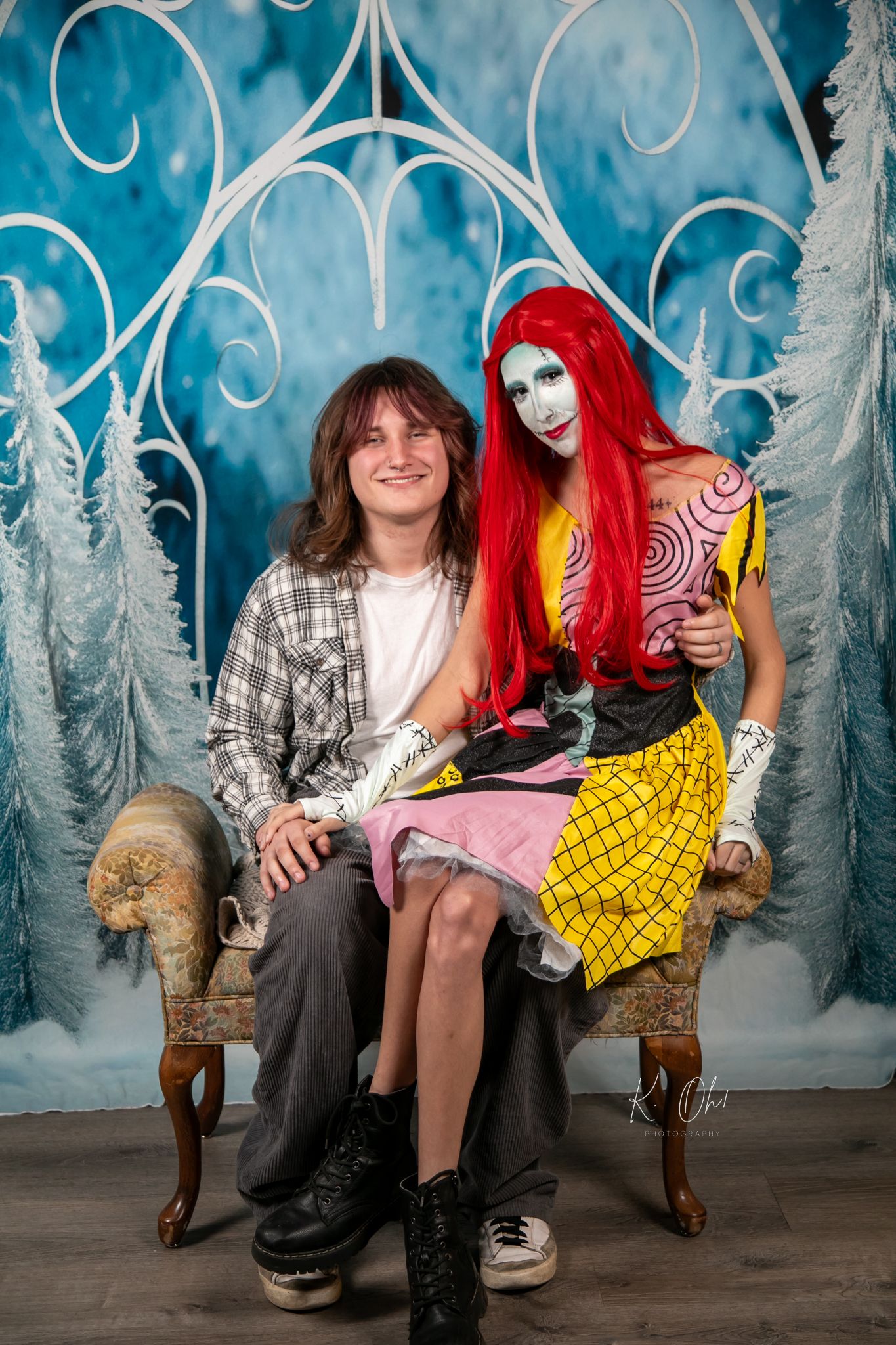 A whimsical winter photo featuring two people sitting on an ornate chair with a magical blue backdrop and white trees.