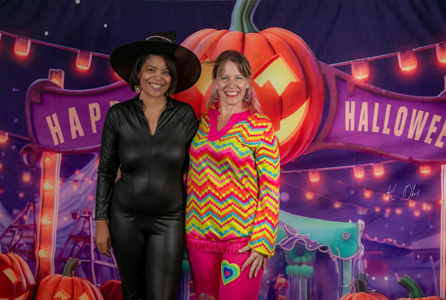 Two people pose in front of a purple Halloween backdrop with glowing jack-o'-lantern decorations.