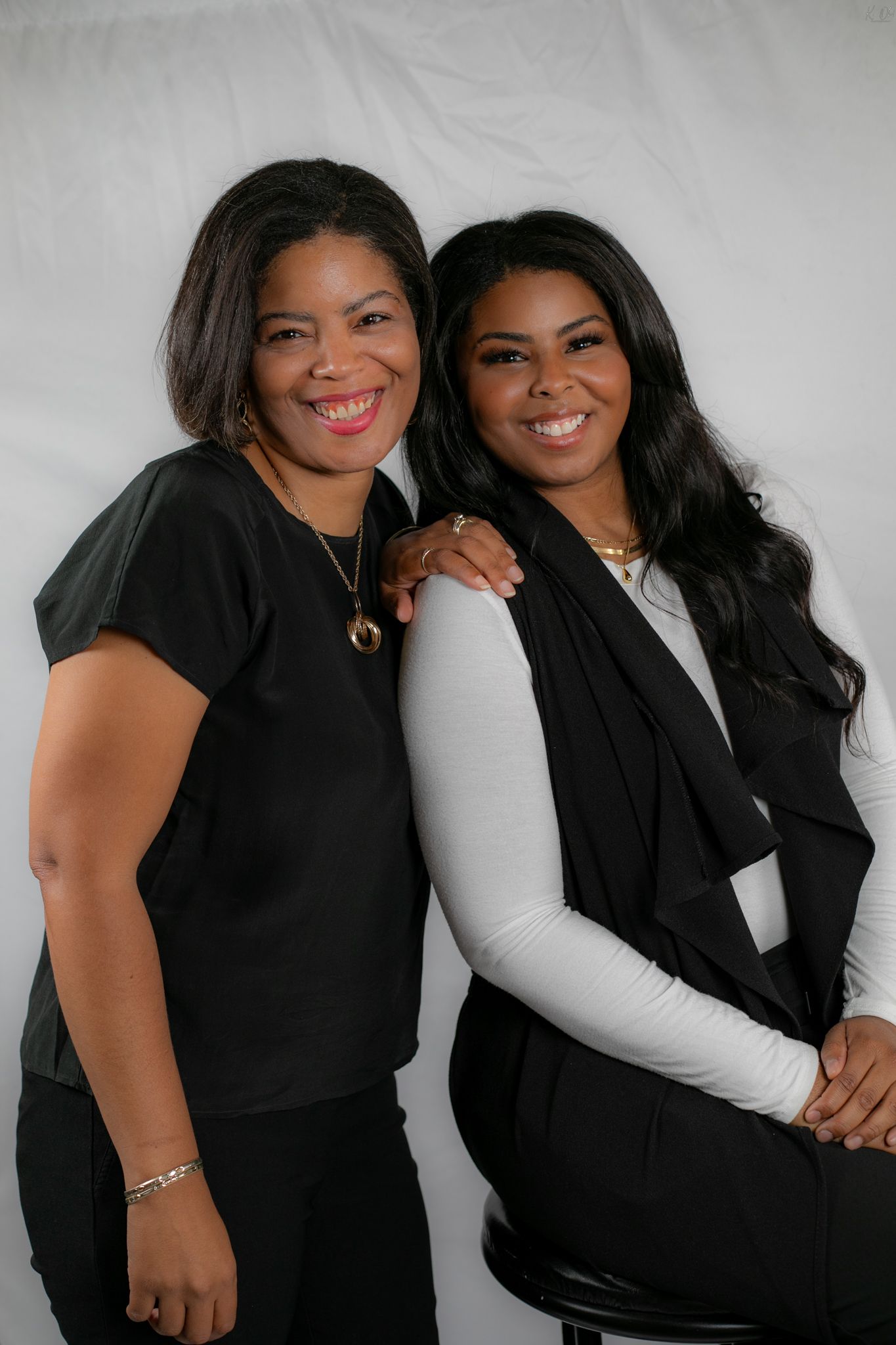 Two women in black outfits posing together for a studio photoshoot against a white background.