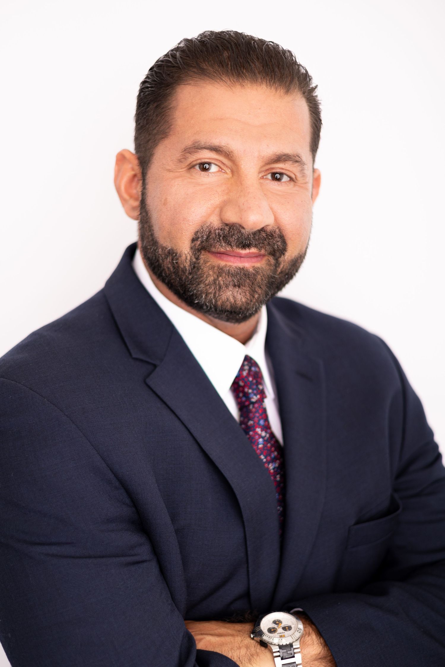 Professional headshot of a bearded man in a navy suit and patterned tie, arms crossed, smiling confidently.