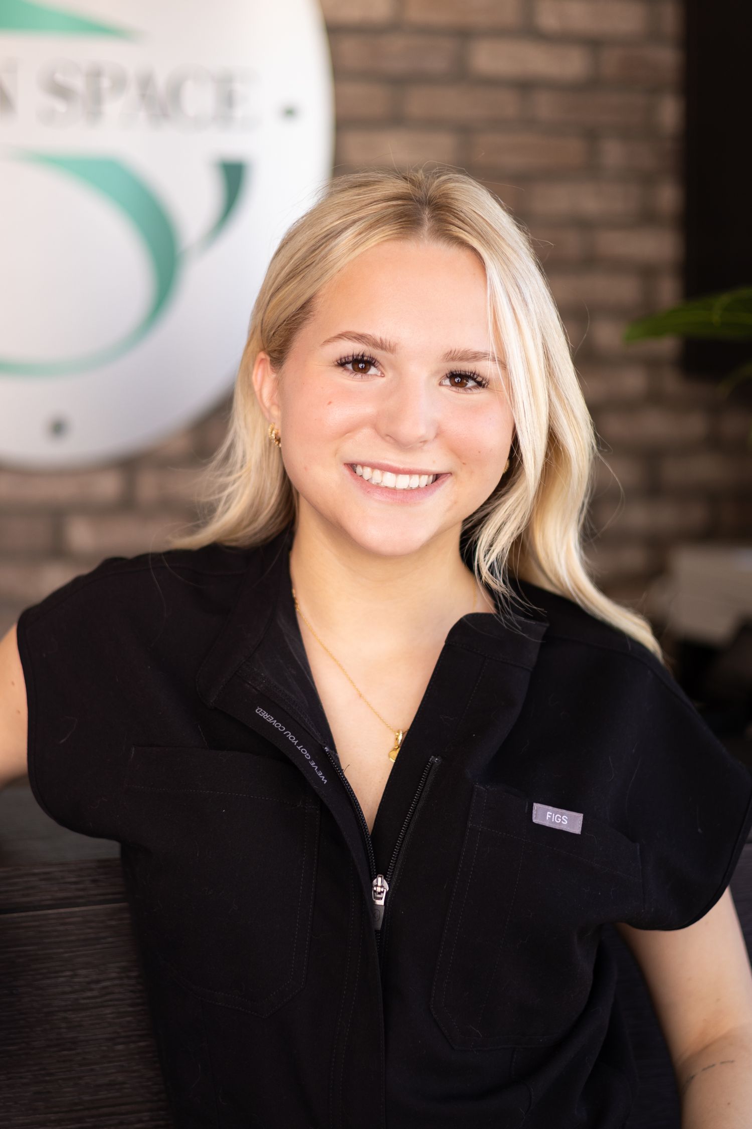 Smiling blonde woman in black scrubs at a modern salon or spa reception area with green logo visible.