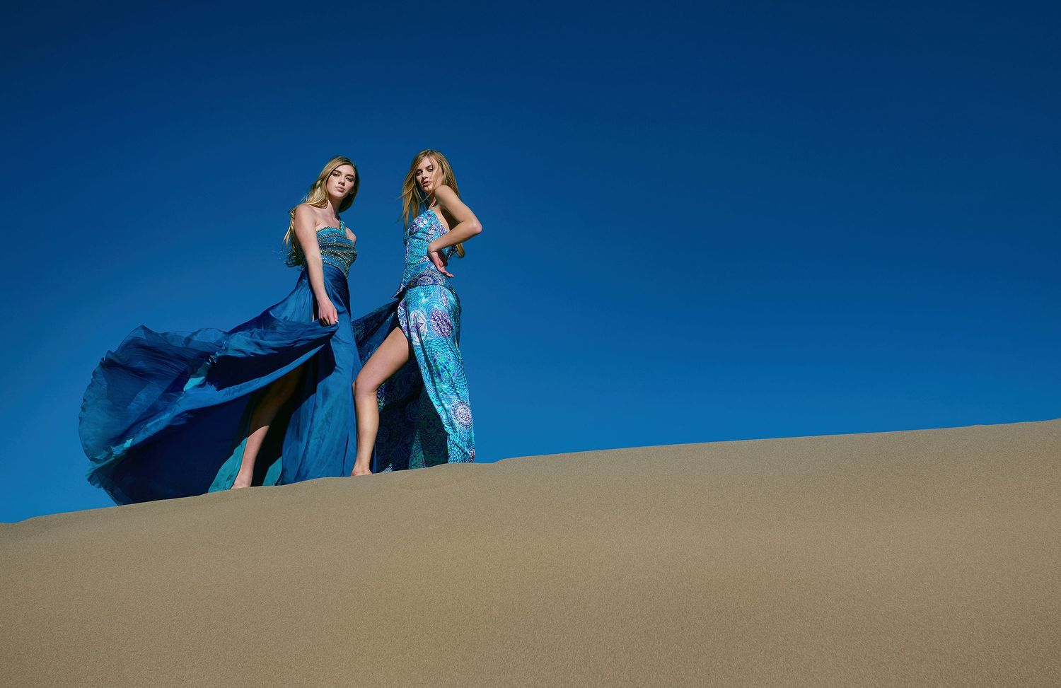 Two women in flowing blue gowns stand dramatically atop a sand dune against a deep blue sky.