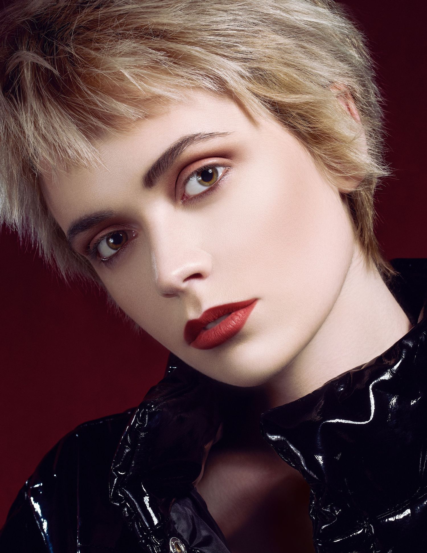 Fashion beauty portrait of model wearing black vinyl top against dark red background with dramatic lighting and bold makeup.
