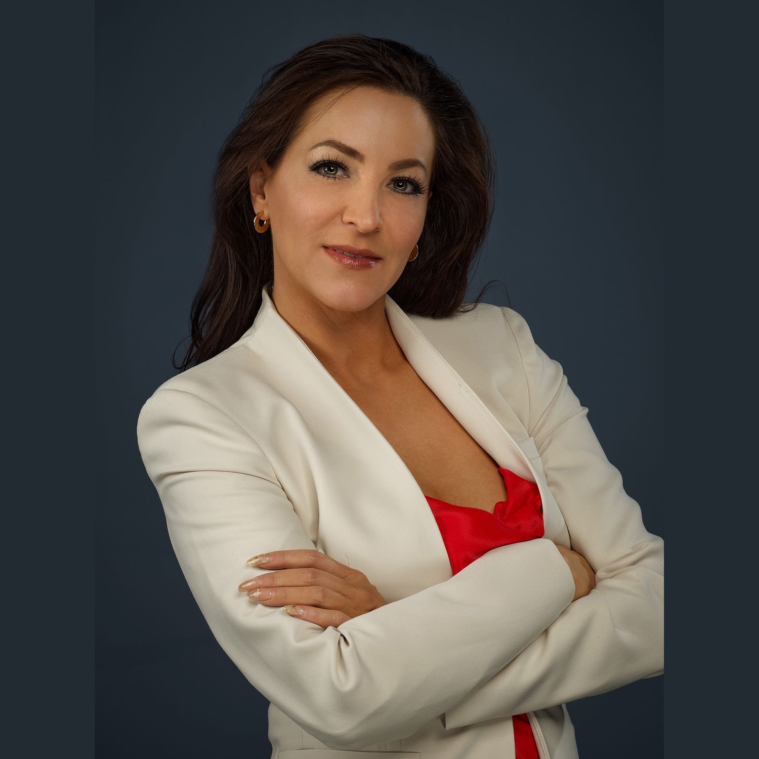 Professional studio portrait of someone in a crisp white blazer and red top posing with arms crossed against dark background.