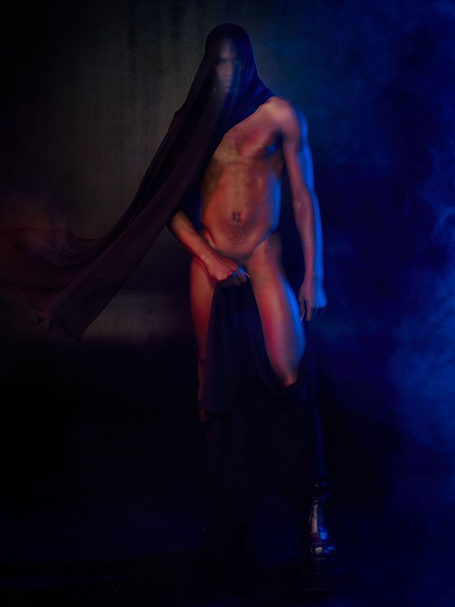Artistic fashion portrait with dramatic blue draping and mask against dark background in moody studio lighting.