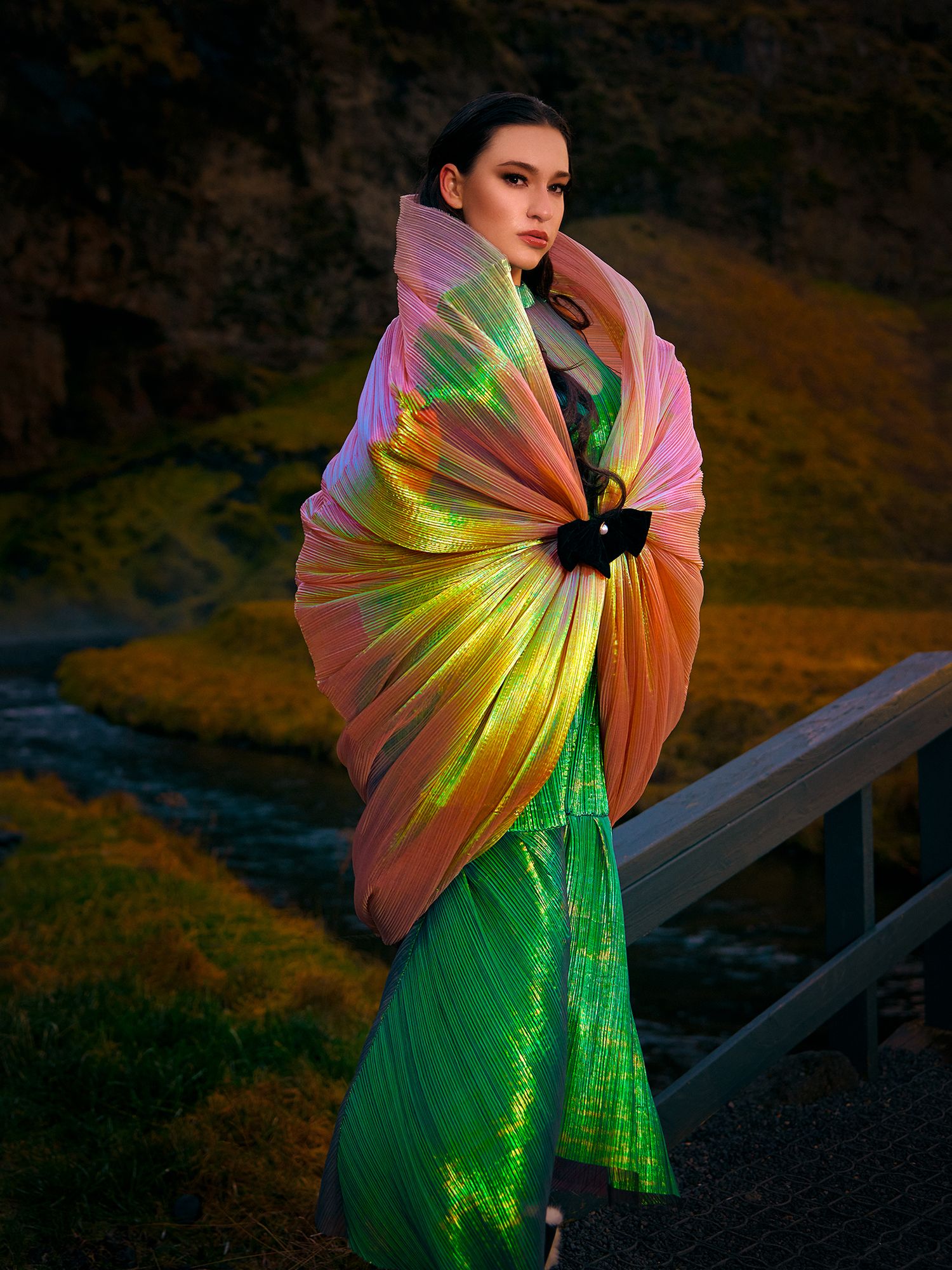 Fashion photography with iridescent shimmering fabric in green and pink hues against a dramatic outdoor background at sunset.