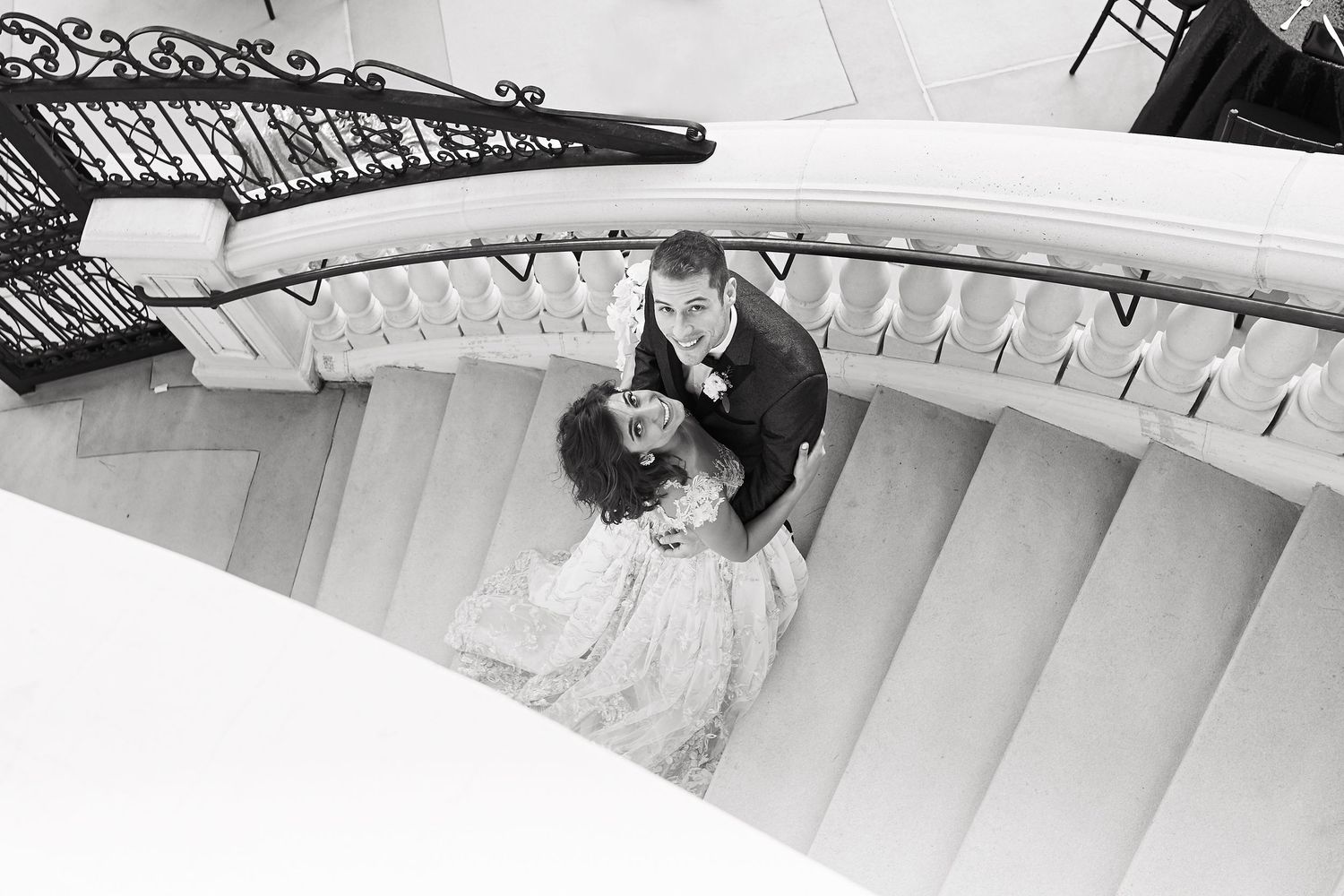 Black and white photograph taken from above showing a couple on an elegant curved white staircase with ornate railings.
