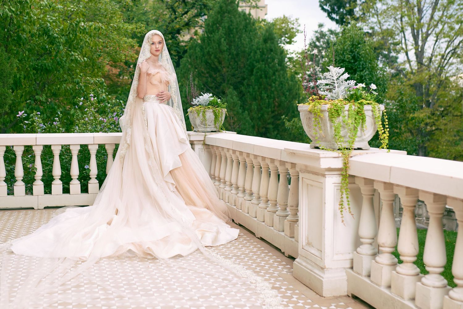 A person in an elegant off-white wedding gown with a flowing train poses on a classic stone balcony with ornate balusters.