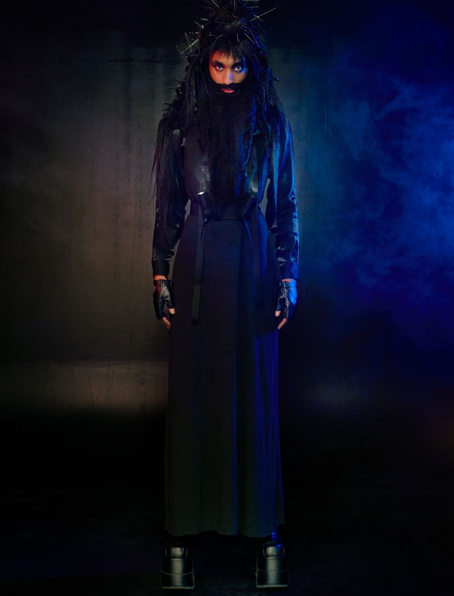 A haunting figure in a long black coat stands on platforms against a dark, moody backdrop with dramatic blue lighting.