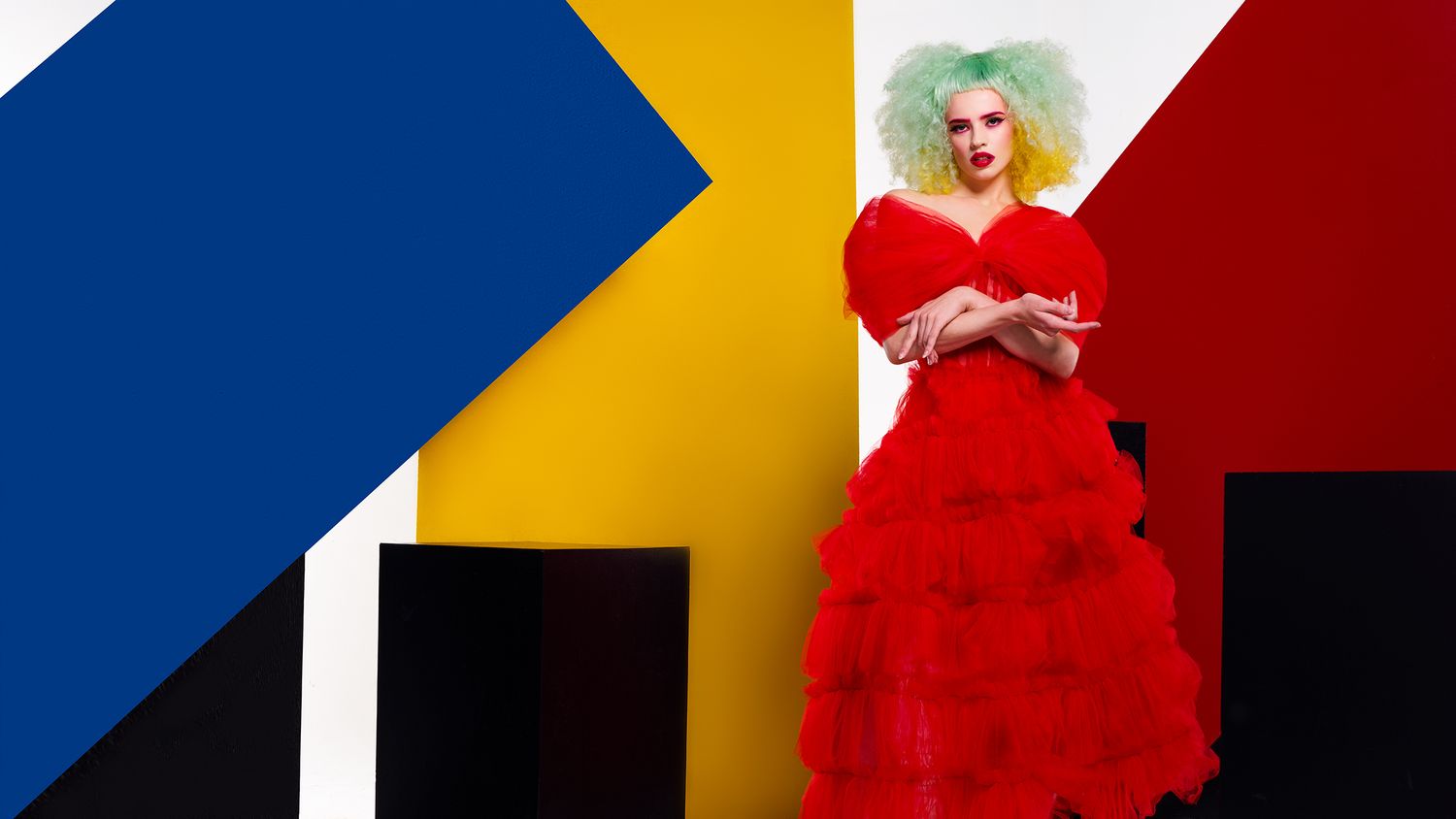 Fashion model in a dramatic red tulle ballgown and colorful hair poses against vibrant geometric color block backdrop.