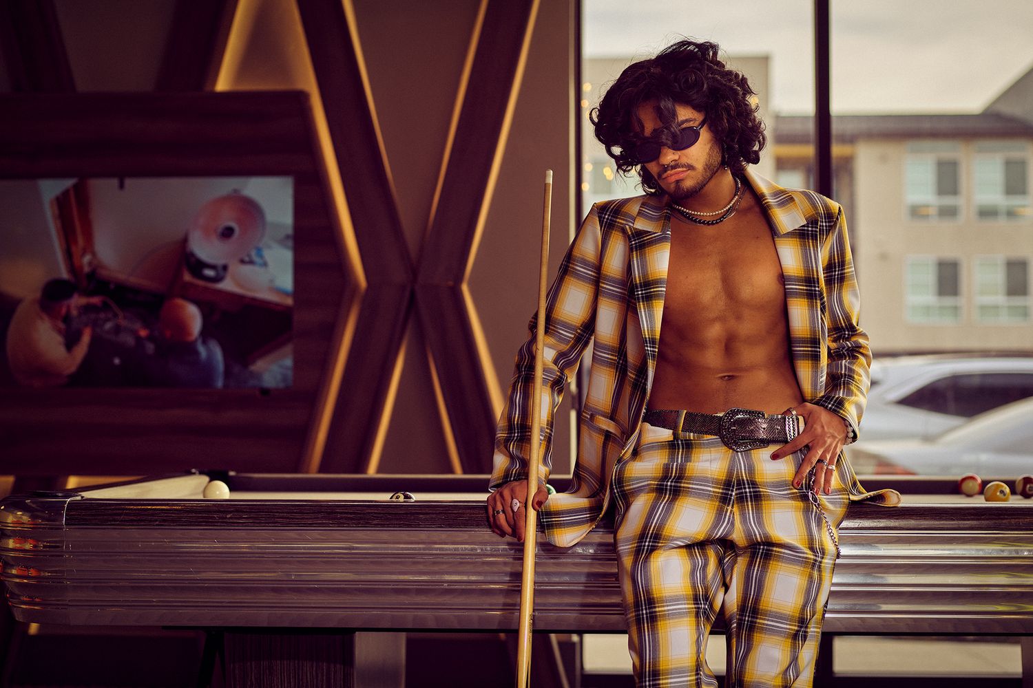 A stylish person leaning on a car wearing a yellow plaid suit and retro-inspired outfit in an urban setting.