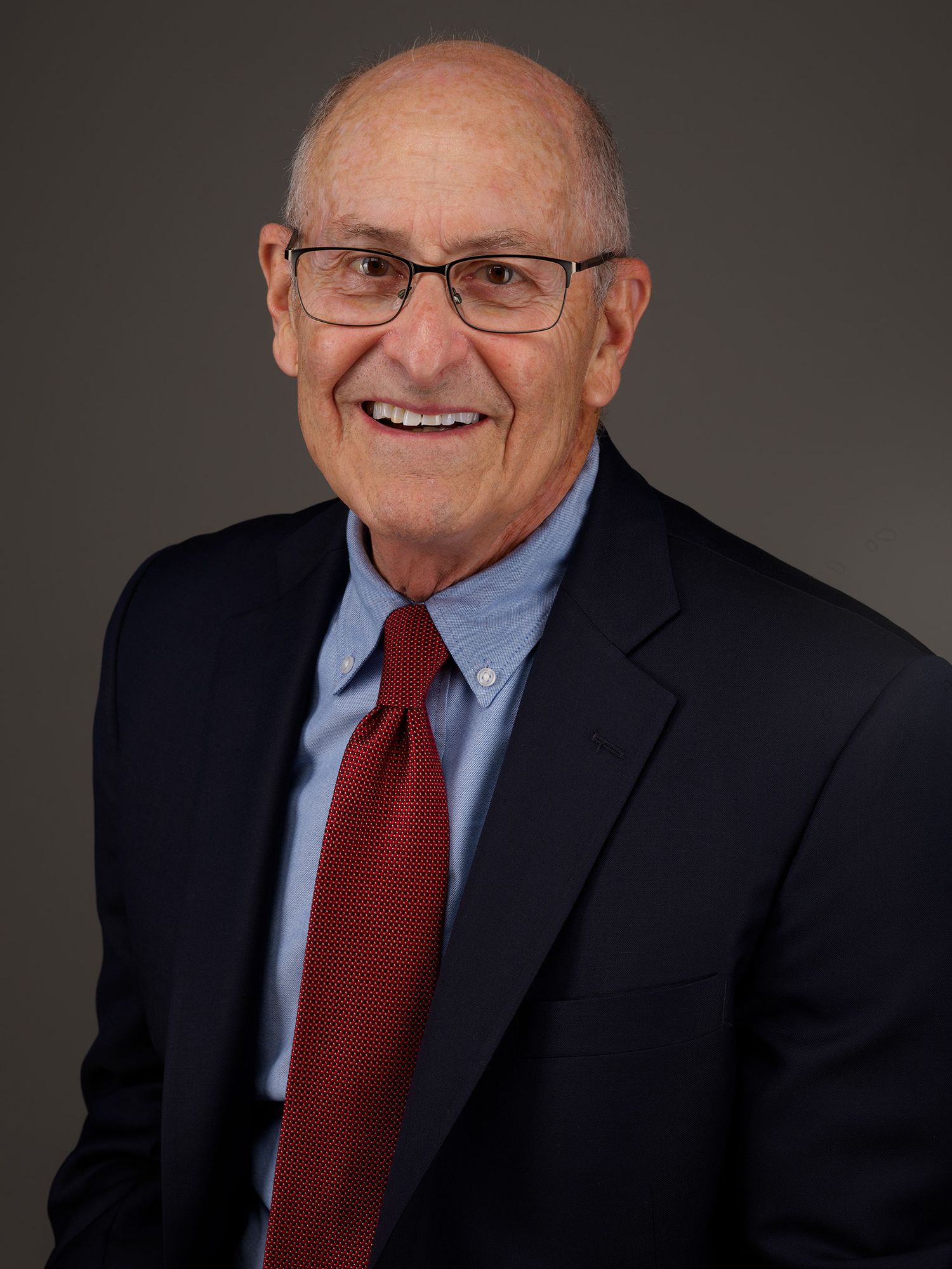 Professional headshot of a senior executive in a navy suit, blue shirt, and red tie smiling warmly against dark background.