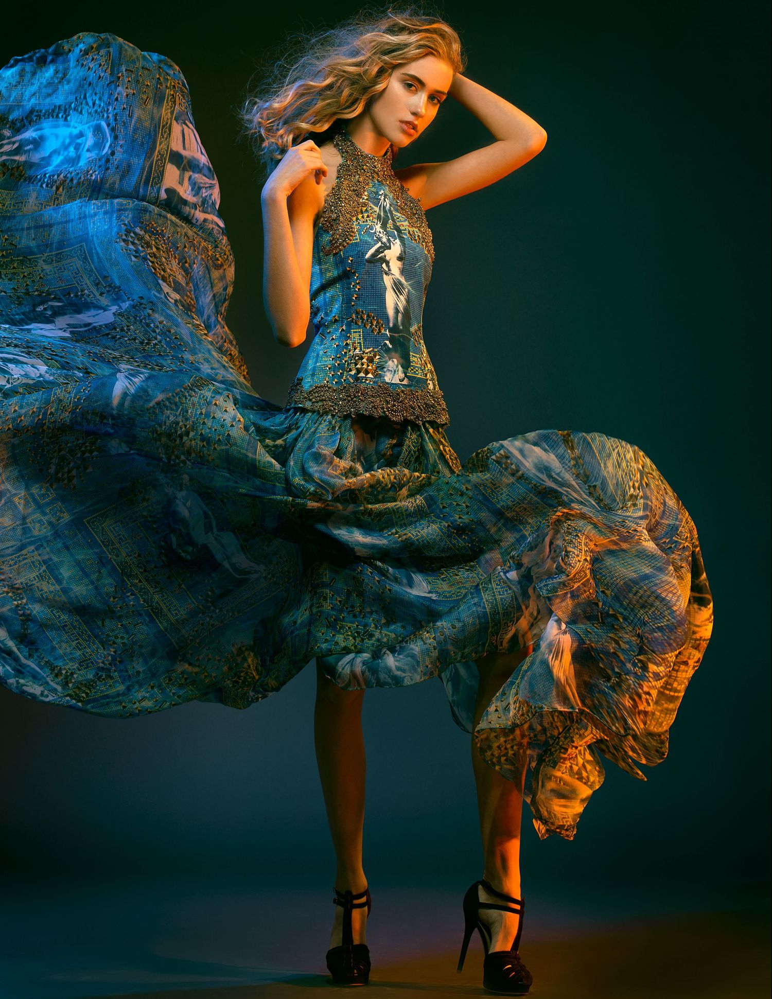 A person in a flowing blue patterned dress poses dramatically against a dark background with atmospheric lighting effects.