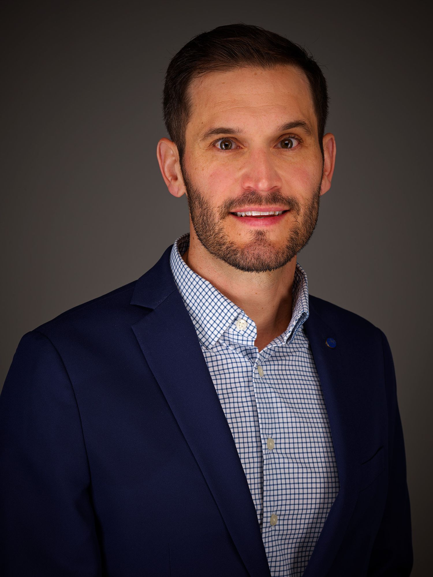 Professional headshot portraits of person wearing navy blazer and blue checkered shirt against dark background.