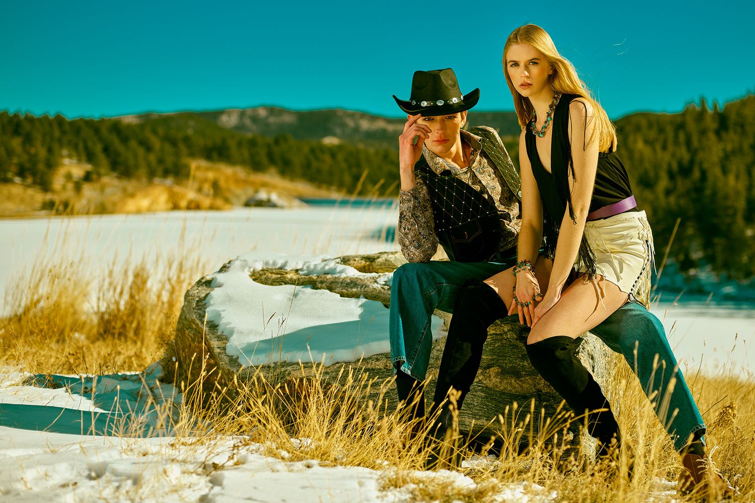 Two individuals in western-style attire pose together against a snowy winter landscape with golden grasses and blue sky.