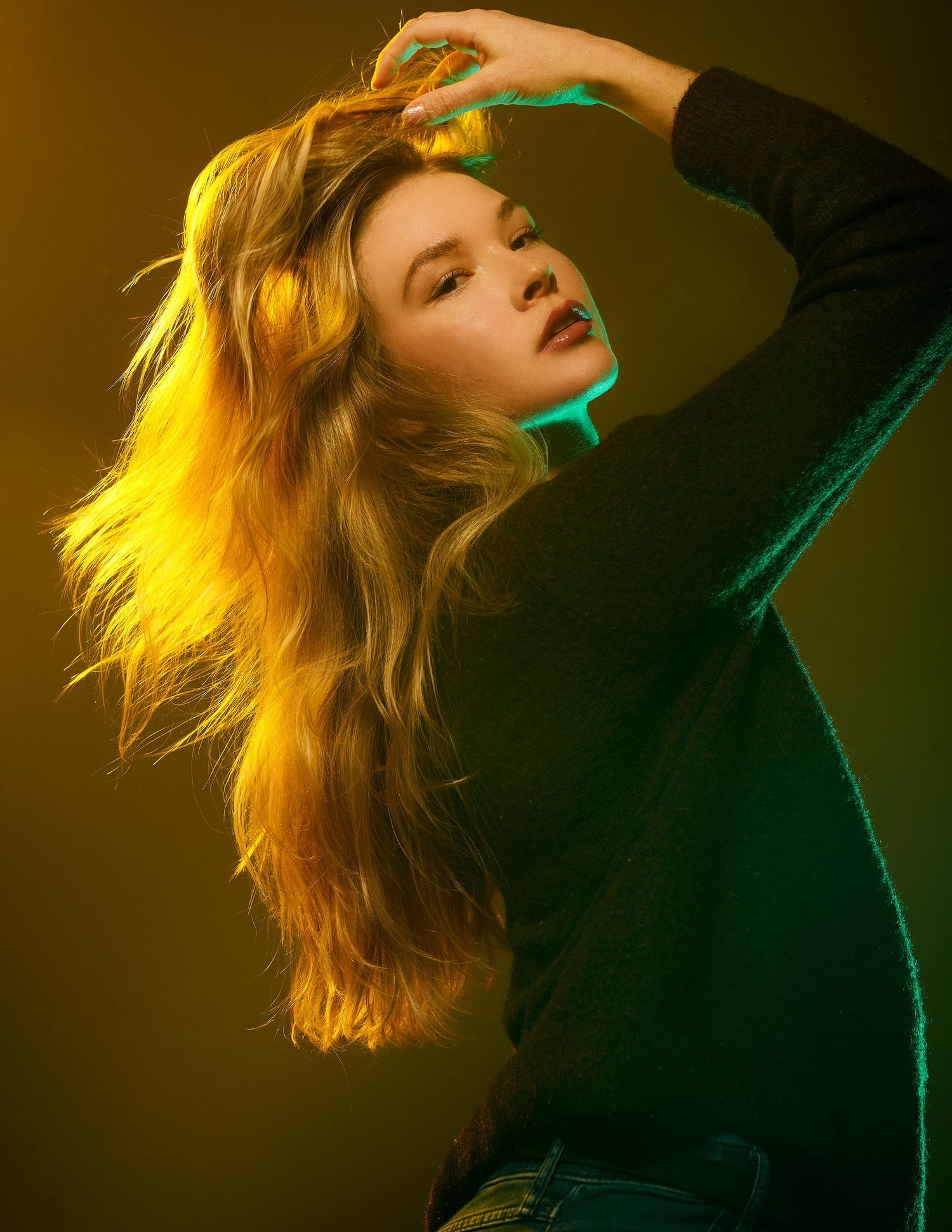 Dramatic fashion portrait with colorful yellow and green lighting casting shadows on long flowing hair and dark outfit.